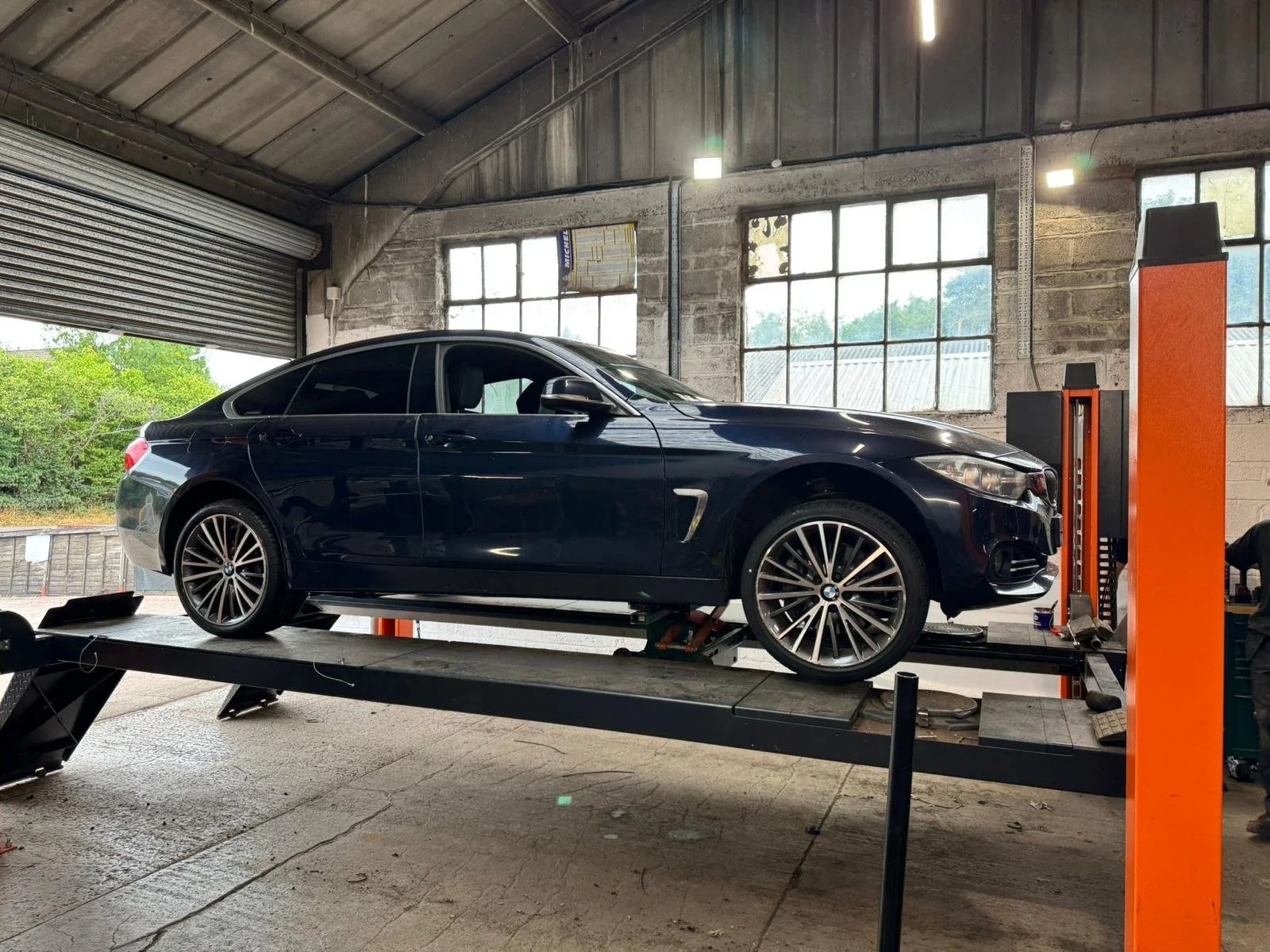 A black BMW sedan on a car lift inside a garage or repair shop, with industrial windows and exposed brick walls.