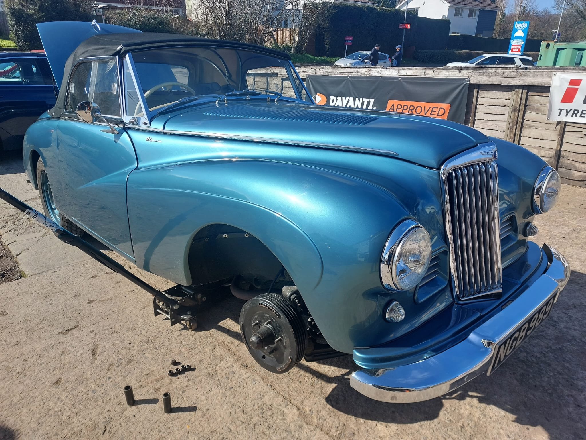 A vintage blue convertible car with its front wheel removed, parked outdoors on a sunny day. The hood is open, and there are various tools and parts lying on the ground nearby. In the background, a wooden barrier with signs, and two people talking, c