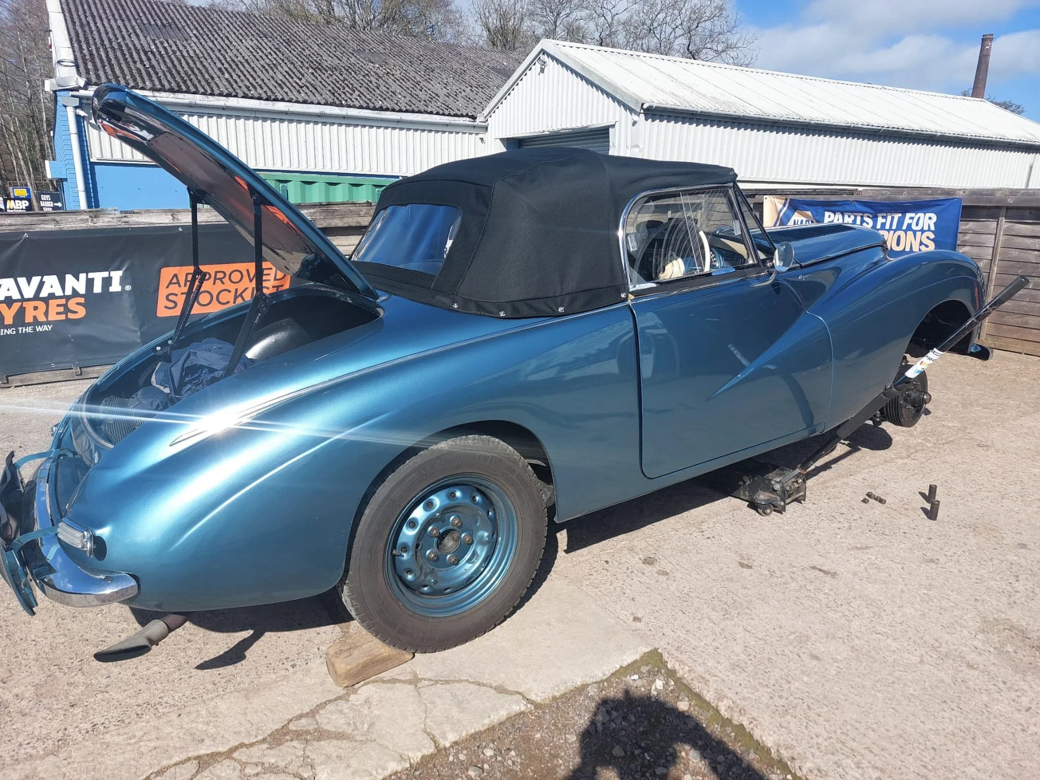 A vintage blue convertible car with a black soft top parked outdoors, with the front wheel resting on a block and the hood open. There are signs and a wooden fence behind the car, and buildings with metal roofs in the background.