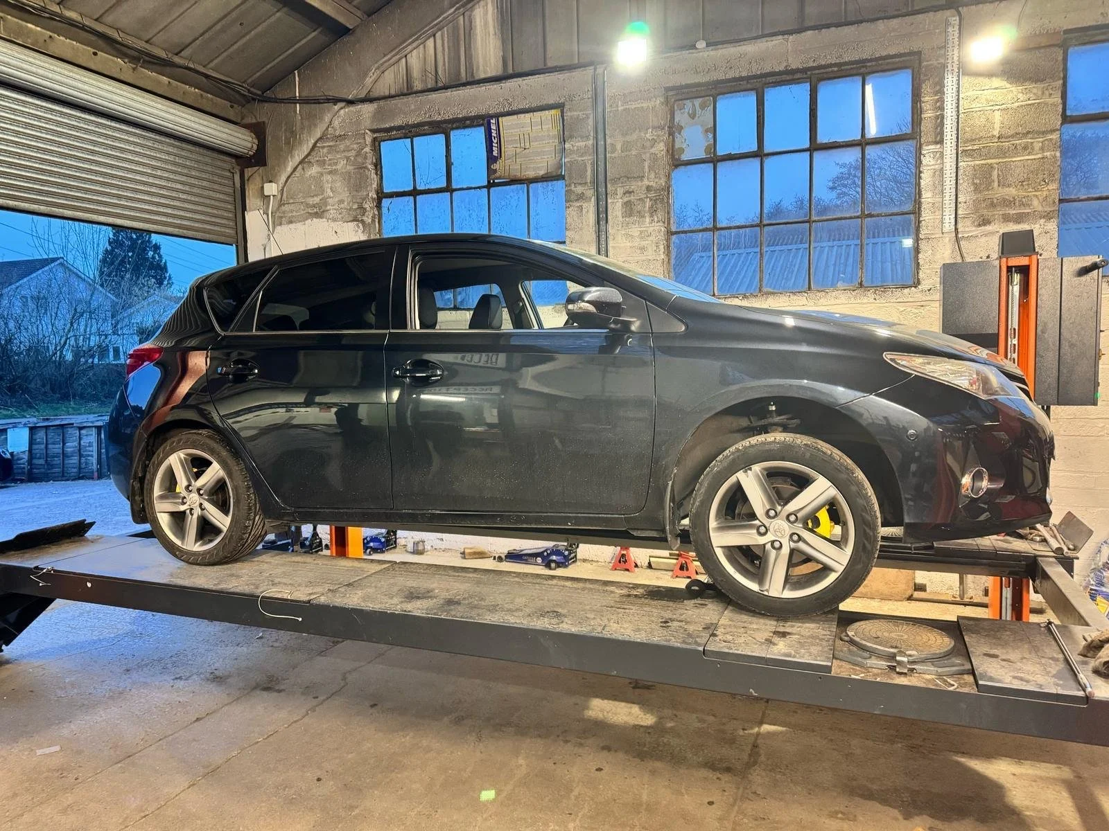 Black car lifted on a hydraulic platform inside an industrial garage with large windows and a roller door.