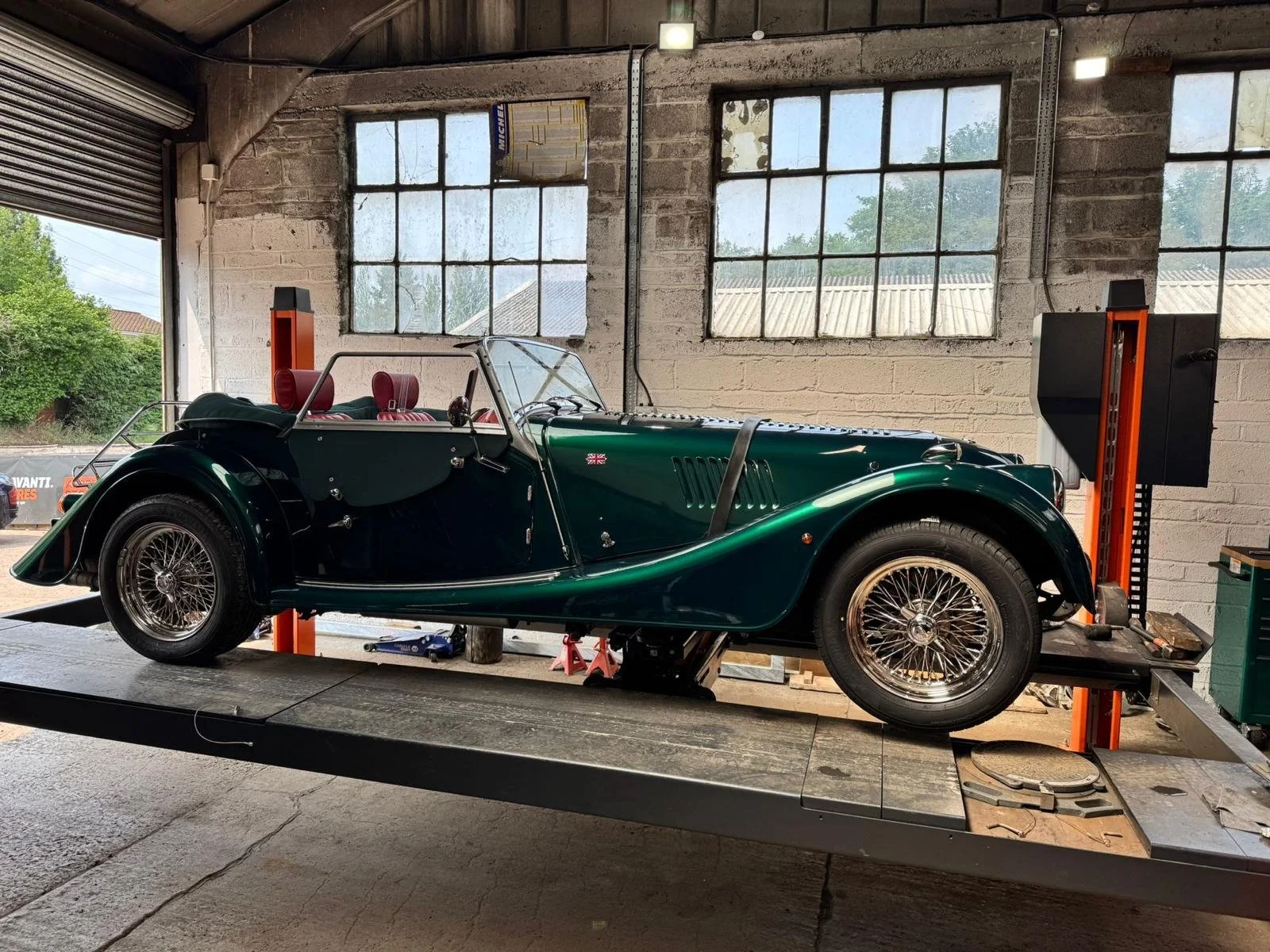 A vintage green convertible car inside a garage, supported by hydraulic lifts, with large industrial windows in the background.