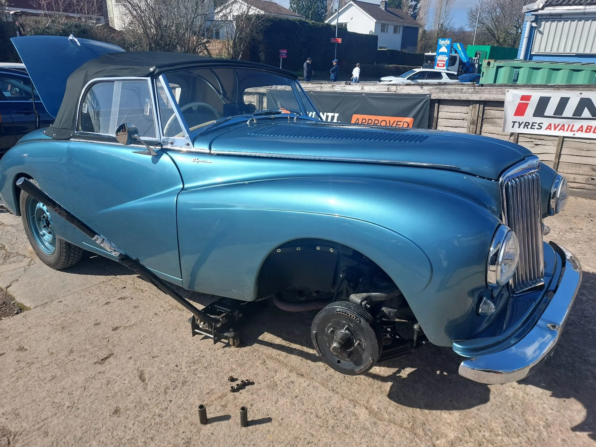 A vintage blue car with a black soft top, missing its front wheel, is parked on a gravel surface at an outdoor car show. In the background, there are people walking, a wooden fence, and signs for tires and Beds Direct.