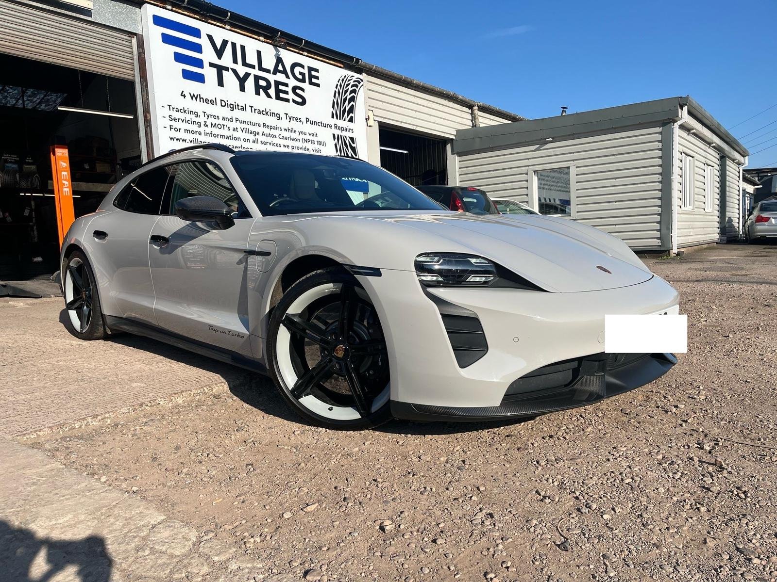 A white Porsche Taycan Turbo electric sports car parked outside of a tire shop with a sign reading 'Village Tyres'.