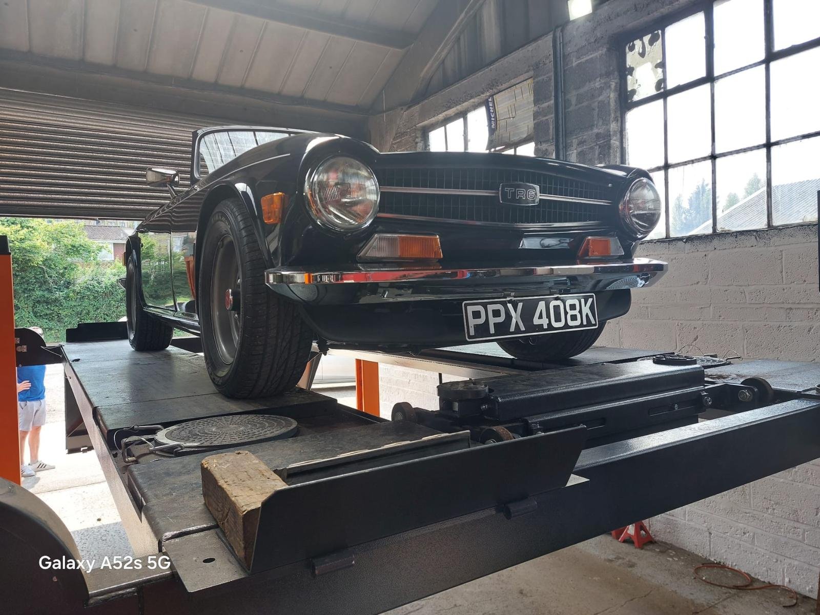 A vintage black sports car on a hydraulic lift inside a garage, with a partially open roller door and a window showing green trees outside.