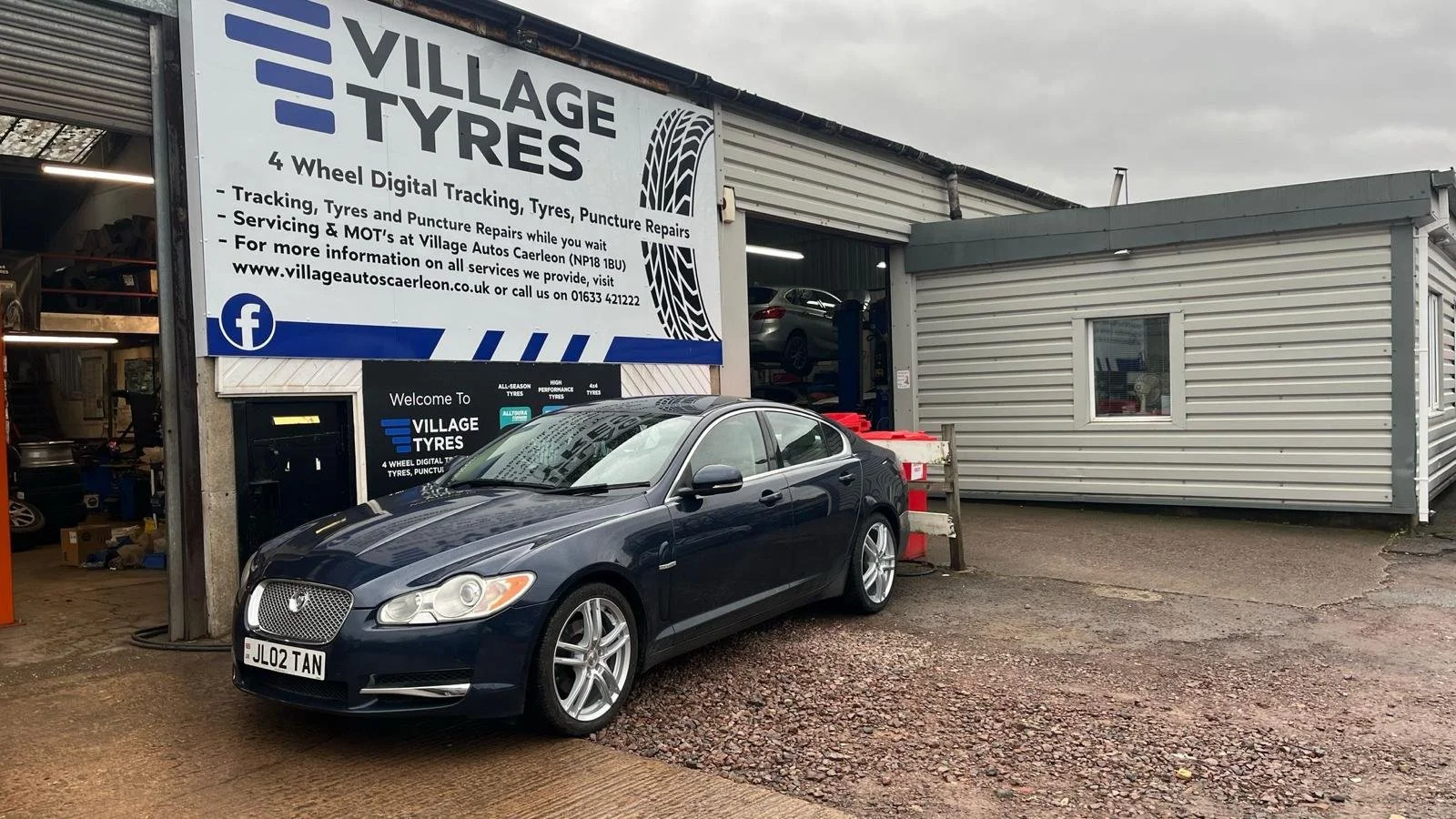 A black sedan car parked outside an auto repair shop called Village Tyres, which has a large sign advertising tire tracking and puncture repairs, with a garage bay in the background showing a car lift and stacked tires.