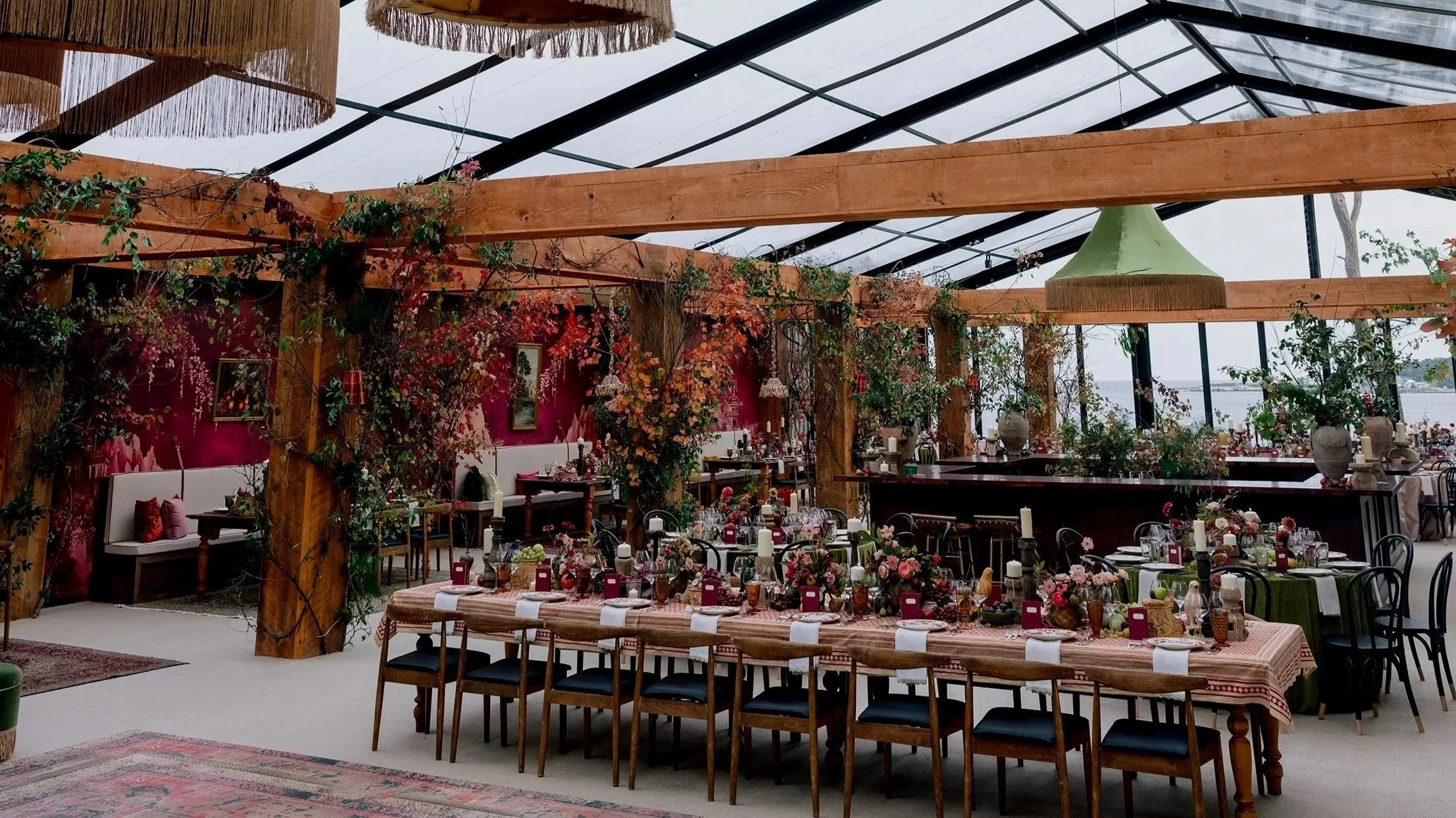 A decorated indoor dining area with long tables set with flowers, candles, and tableware, surrounded by wooden beams and green plants, with large windows showing an overcast sky outside.