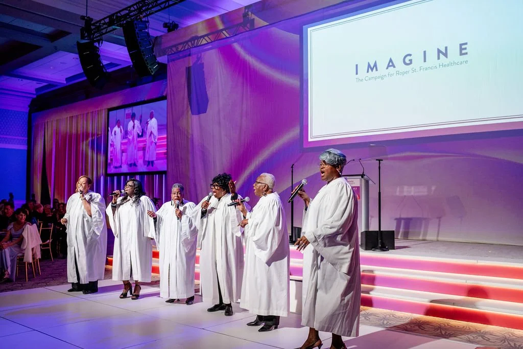 Group of women dressed in white choir robes singing on stage at a church or conference event, with large screen displaying 'IMAGINE' and the subtitle 'The Campaign for Roper St. Francis Healthcare.' Audience seated on the left.