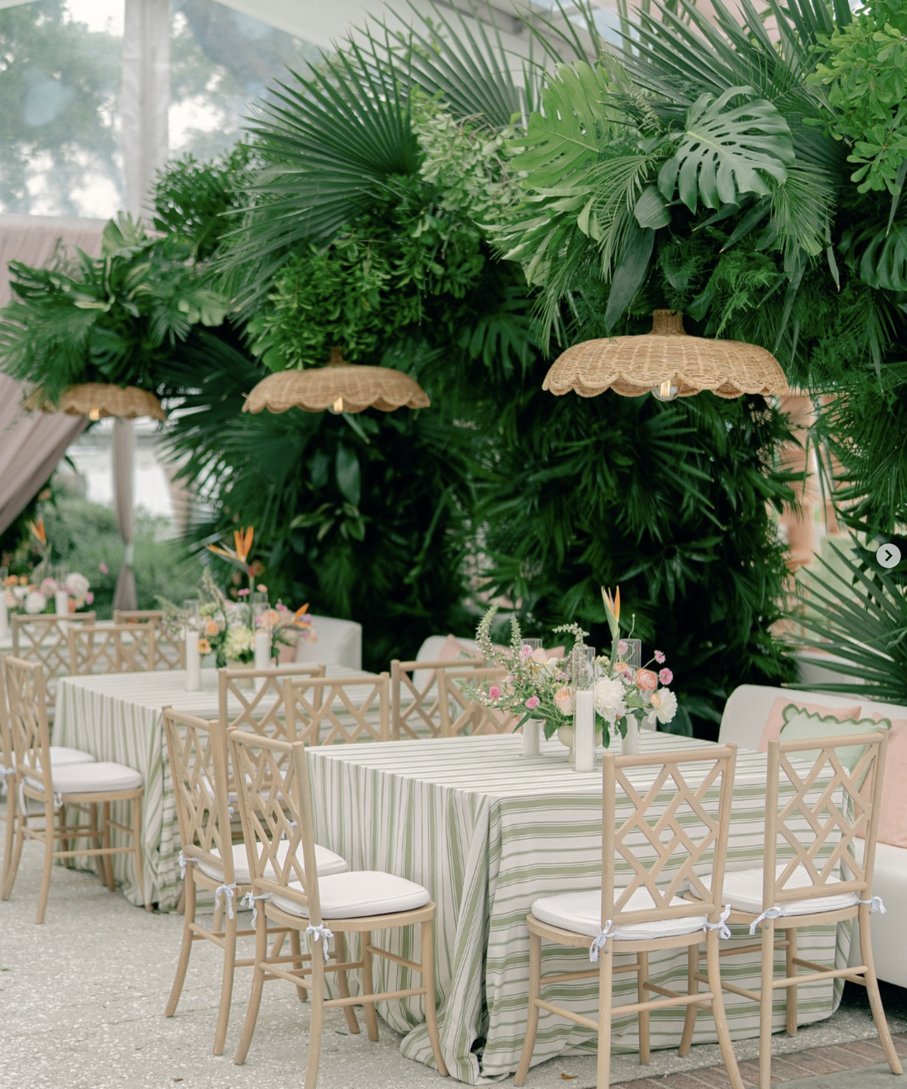 Round tables with striped tablecloths, surrounded by wooden chairs with white cushions, set up for an outdoor event with floral centerpieces, under hanging woven lamps, with tropical plants as a backdrop.