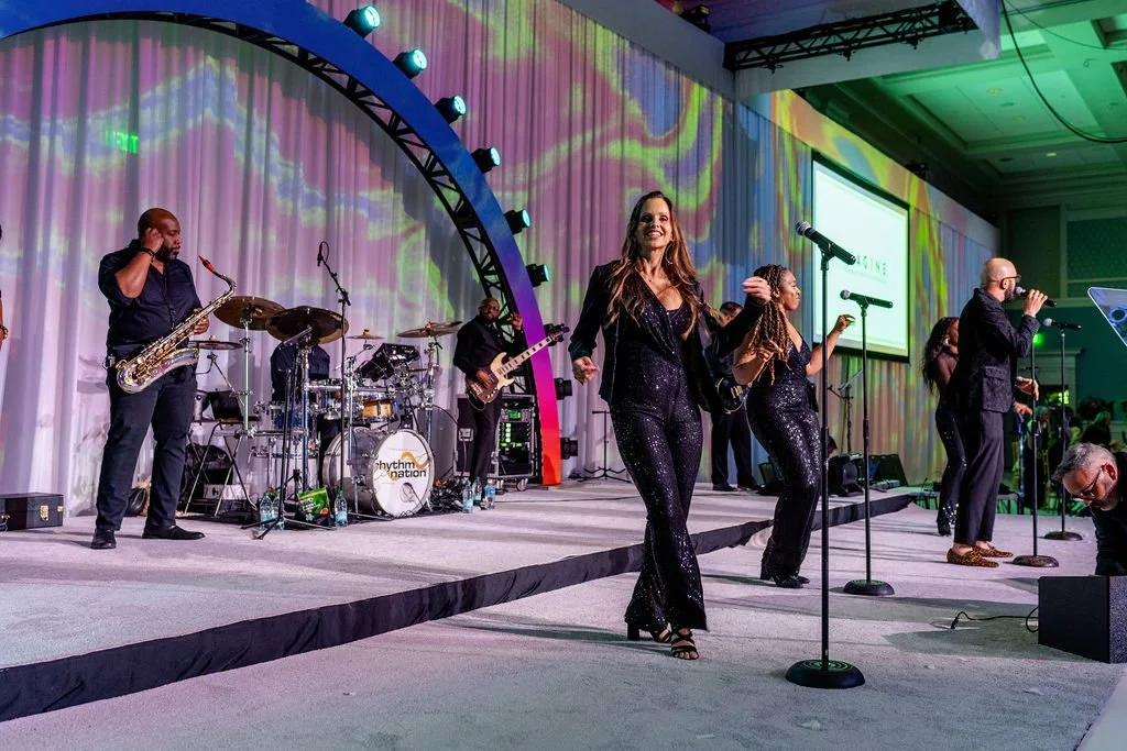 A live musical performance with a band on stage, including singers and musicians, in front of a colorful, lighted backdrop at an event or party.