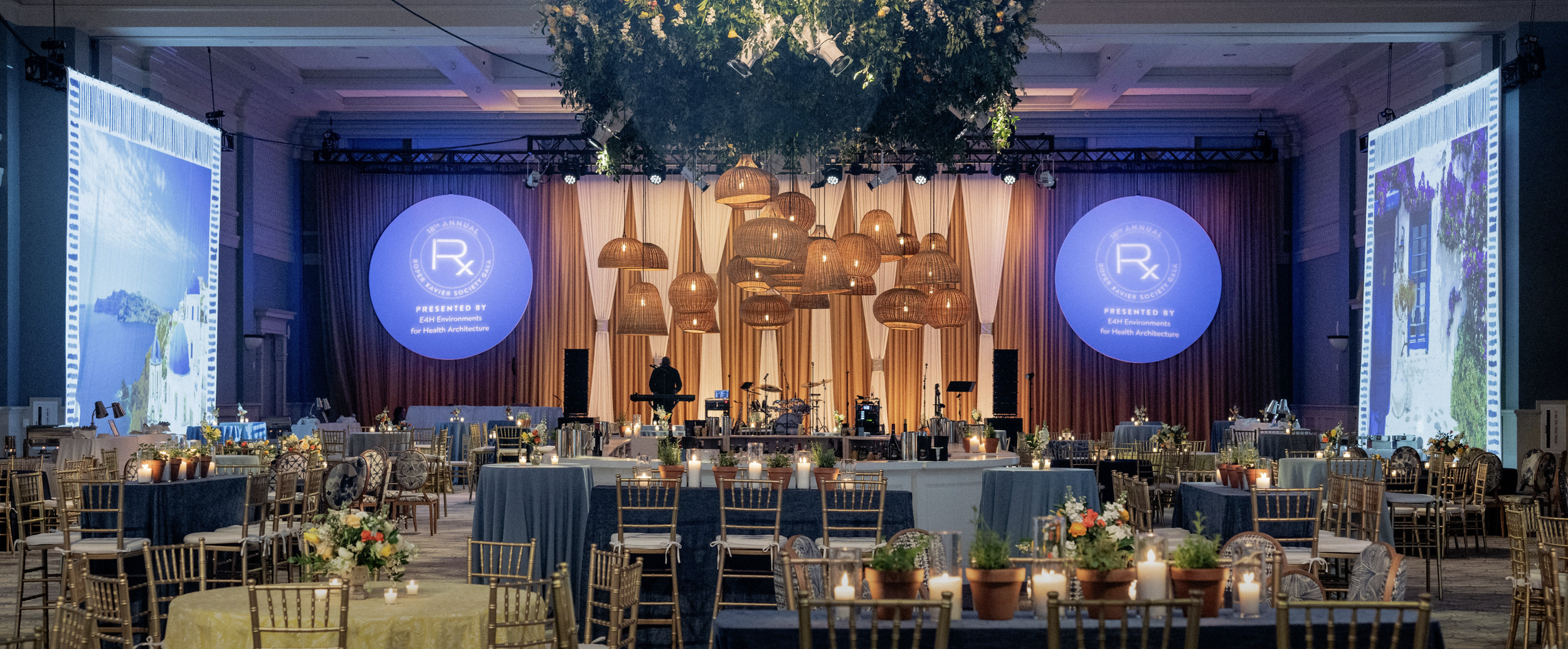 A large event hall decorated with hanging woven pendant lights, round tables with floral centerpieces and candles, and a stage with large screens displaying the Rx symbol and event details.