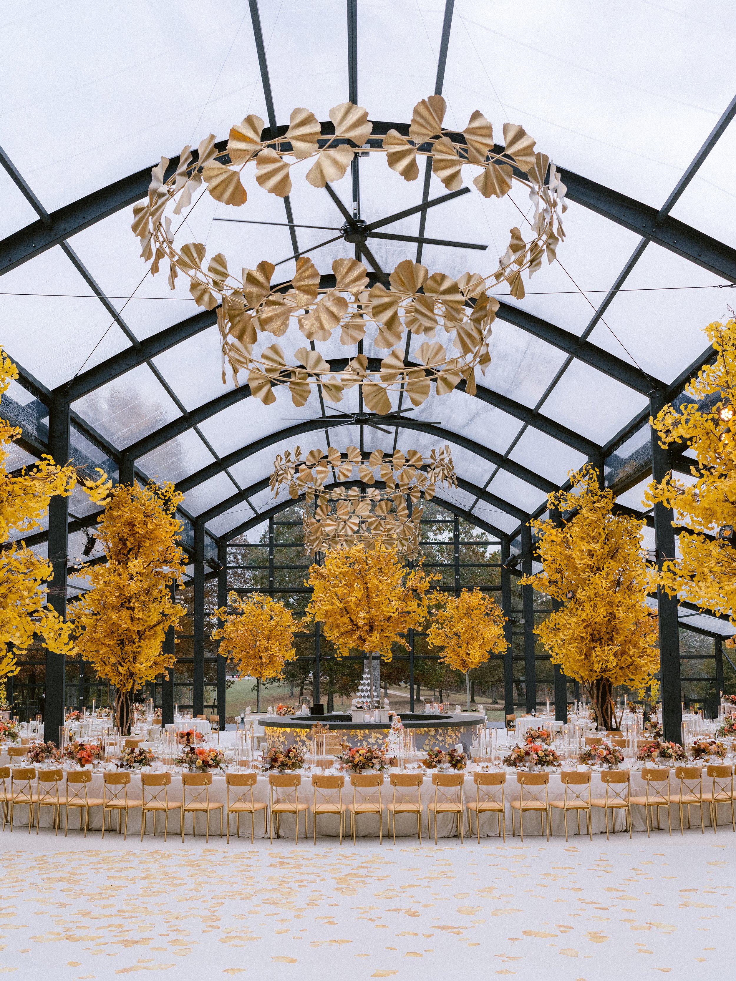 A decorated indoor wedding reception area with a high arched ceiling made of glass panels, featuring a large circular chandelier adorned with gold-colored ginkgo leaves and tall trees with yellow leaves, with round tables set with flowers and chairs arranged around the room.