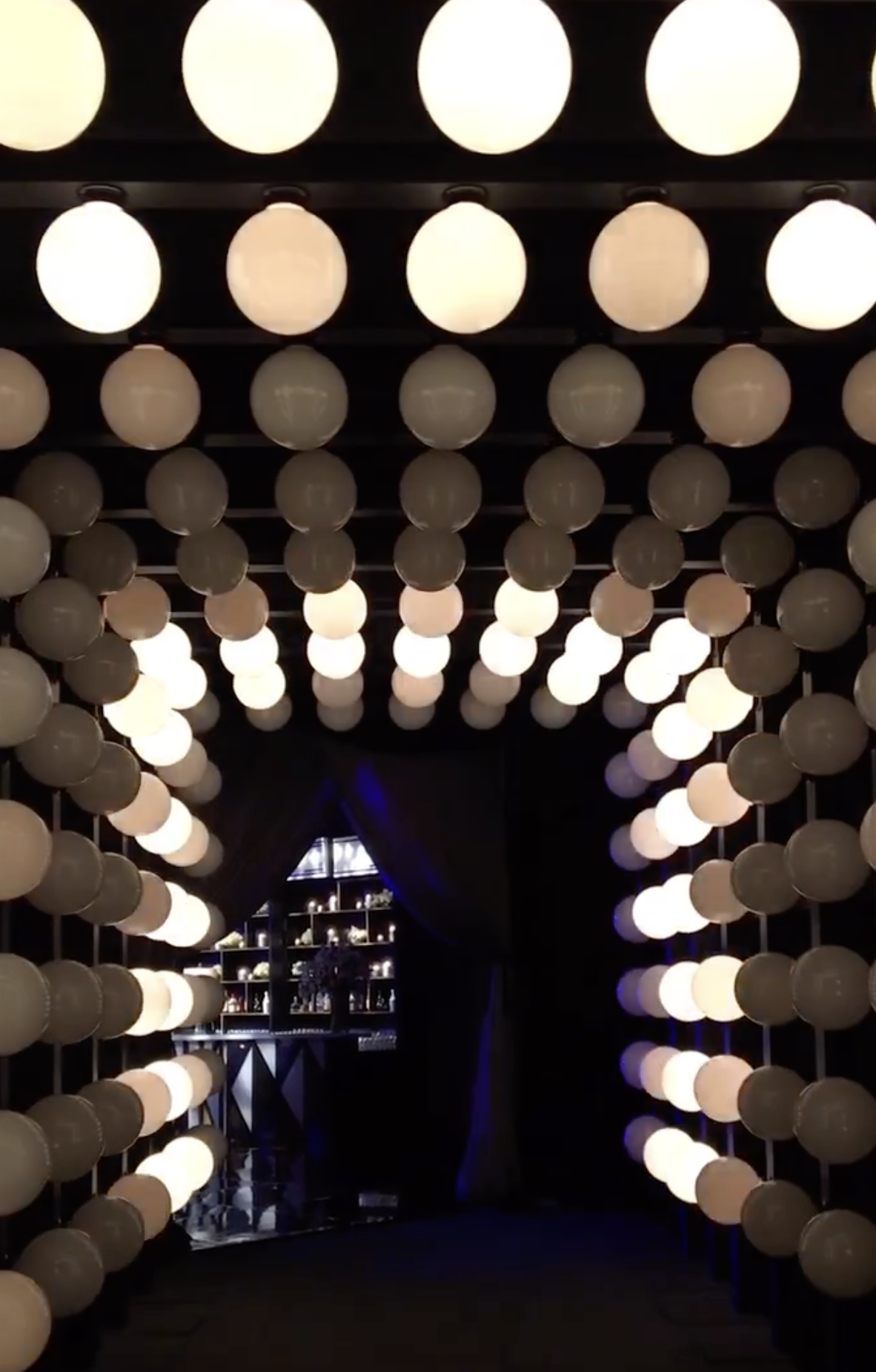 A dark hallway decorated with multiple rows of glowing white spherical lights hanging from the ceiling, leading to an area with a shelving unit and dark curtains.