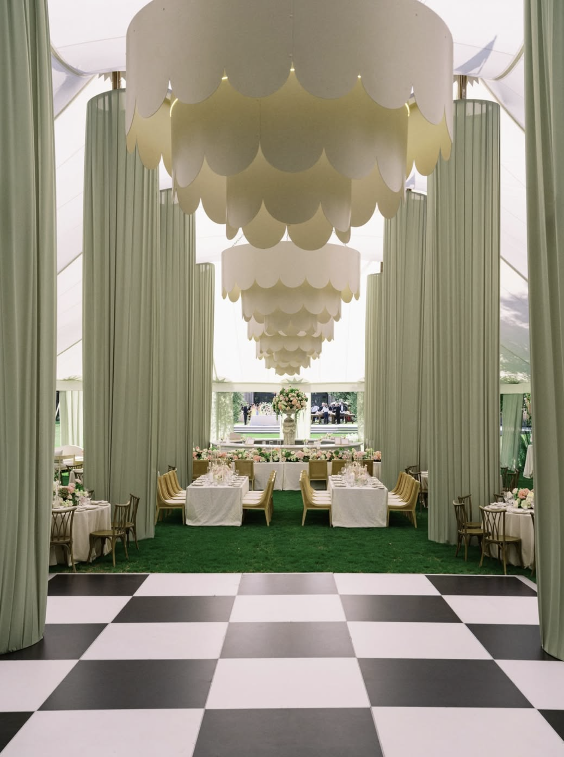 Indoor wedding reception scene with black and white checkered floor leading to a decorated space with long tables, floral centerpieces, and a large tiered chandelier hanging from the ceiling.