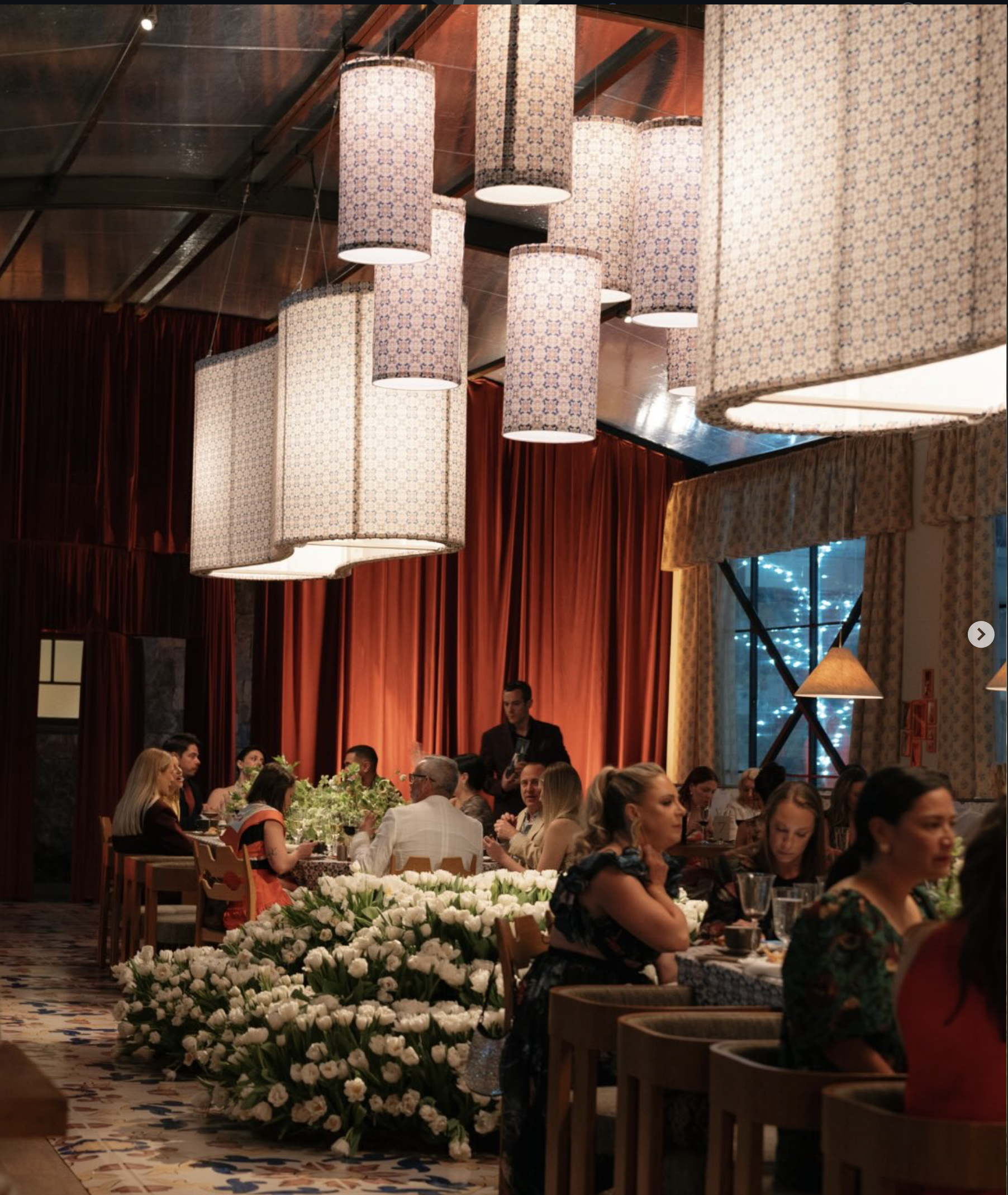People dining at a formal event with large floral arrangements on the tables, warm lighting, and elegant interior decor including hanging patterned lamps and drapes.