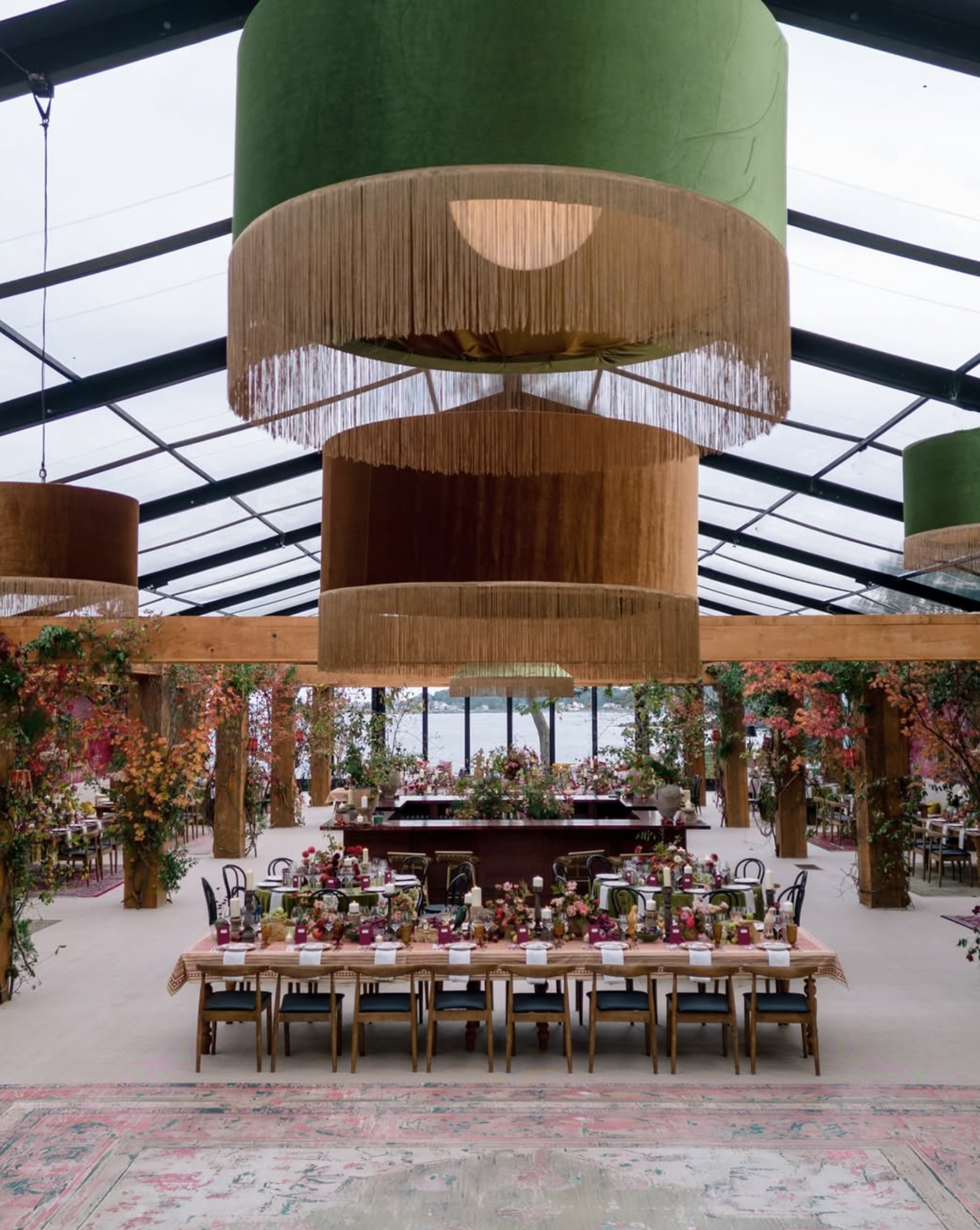 Event space decorated with floral arrangements and table settings, under a ceiling with large hanging light fixtures and wooden beams in a bright, airy room with floor-to-ceiling windows overlooking a body of water.