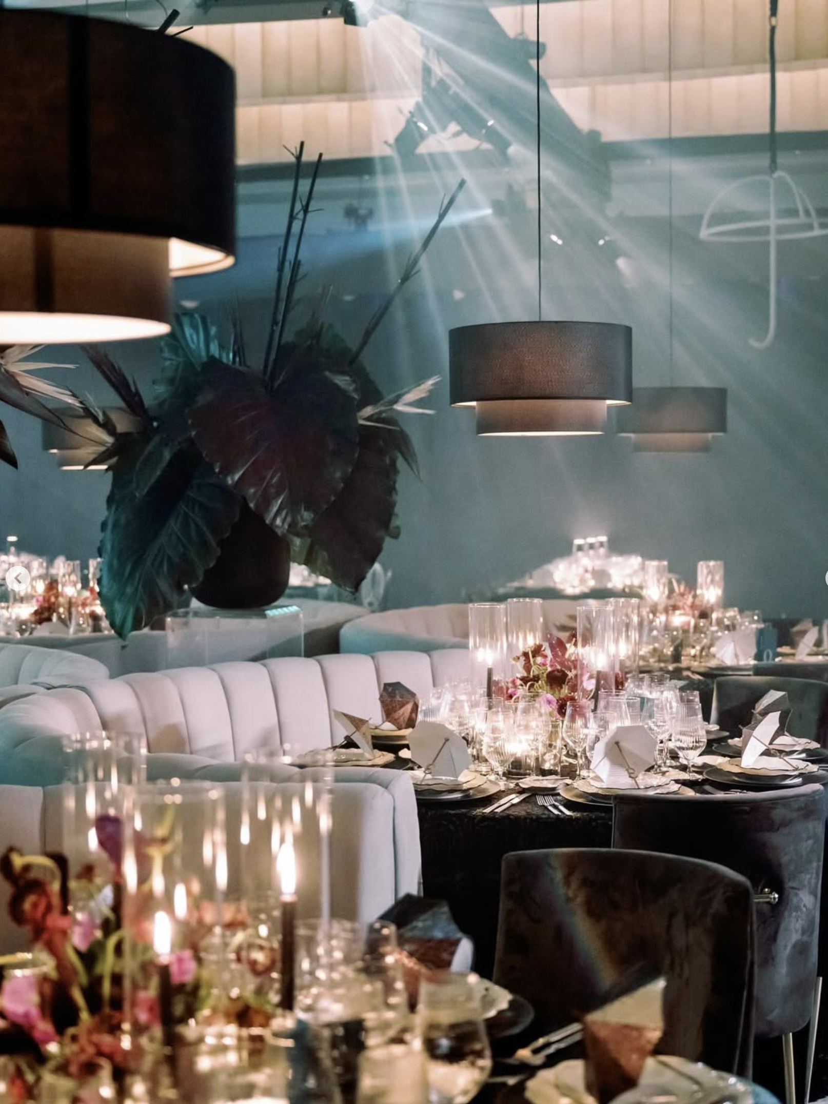 Formal dining area with tables set for an event, featuring candles, glassware, and floral arrangements; modern decor with hanging pendant lights and a large potted plant.