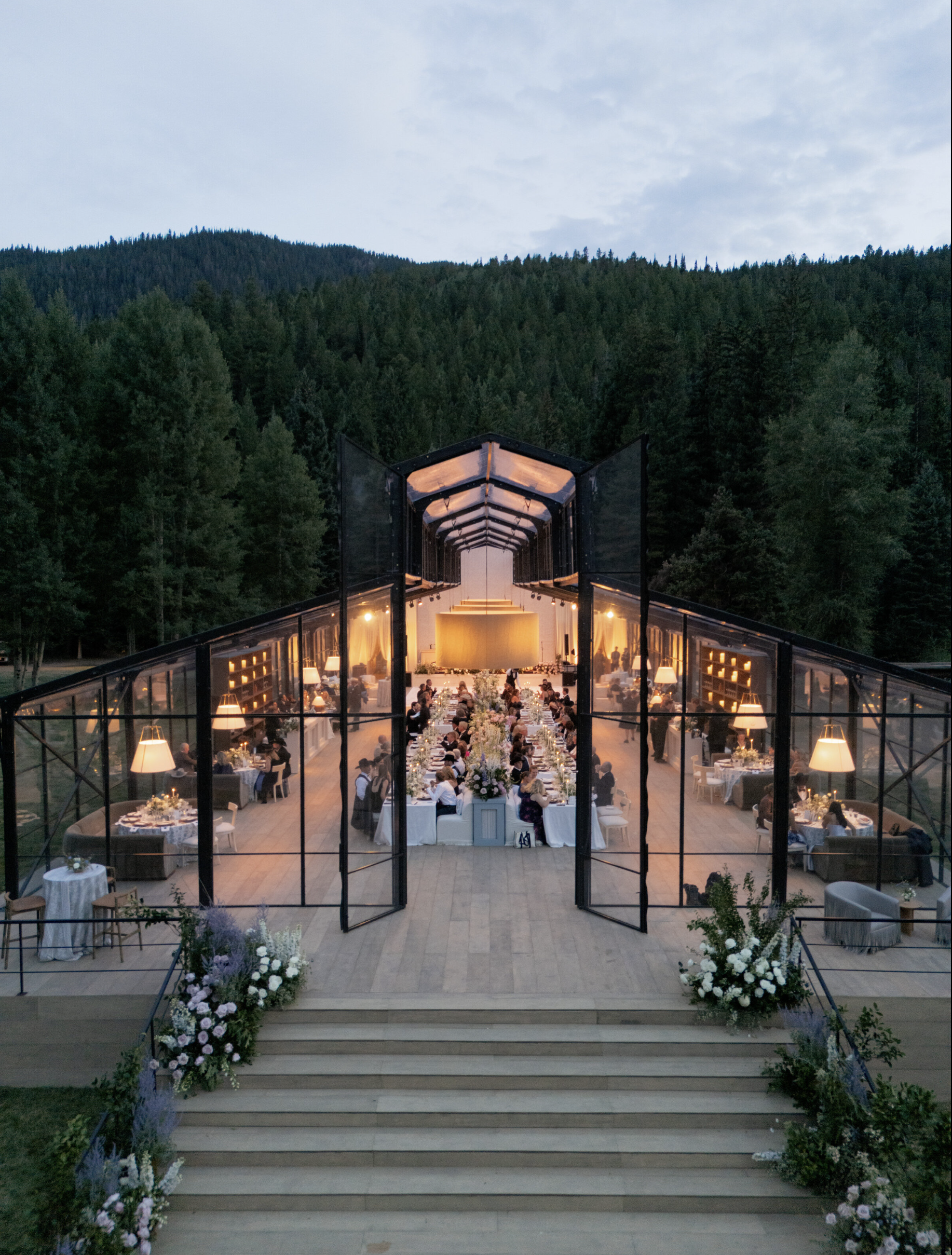 A large glass-walled wedding reception inside a pavilion surrounded by forested mountains at dusk. Decorated tables with white tablecloths, floral centerpieces, and guests seated, with a view of the outdoor steps decorated with flowers leading up to the pavilion.