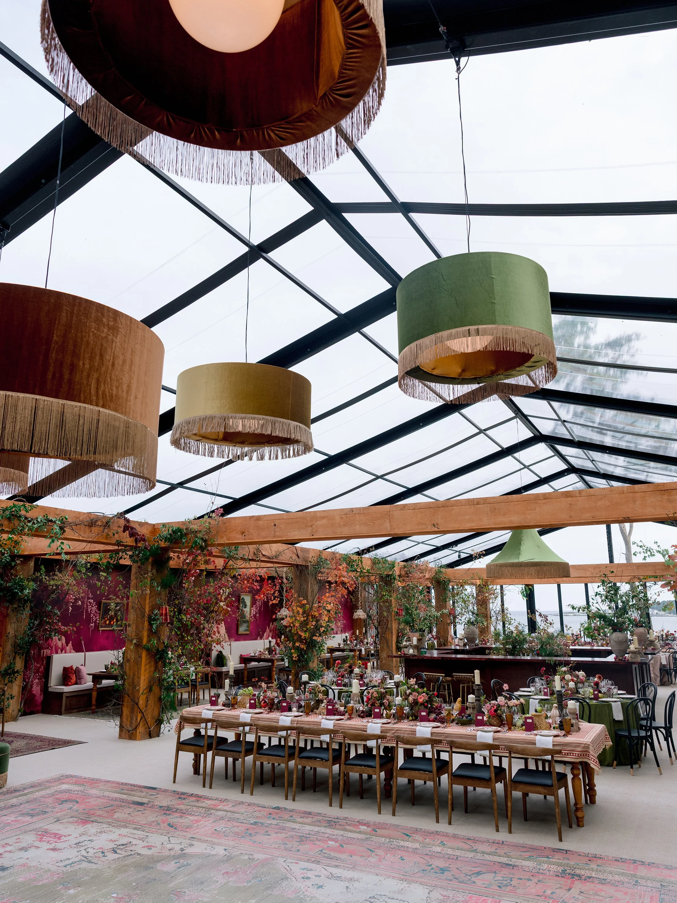 A decorated indoor dining space with tables set for an event, featuring floral arrangements, candles, and purple menus. Large colorful hanging lampshades are suspended from the ceiling, which is made of glass beams, creating a bright, airy atmosphere. The walls are painted pink with floral decorations, and the space is furnished with wooden and metal chairs.