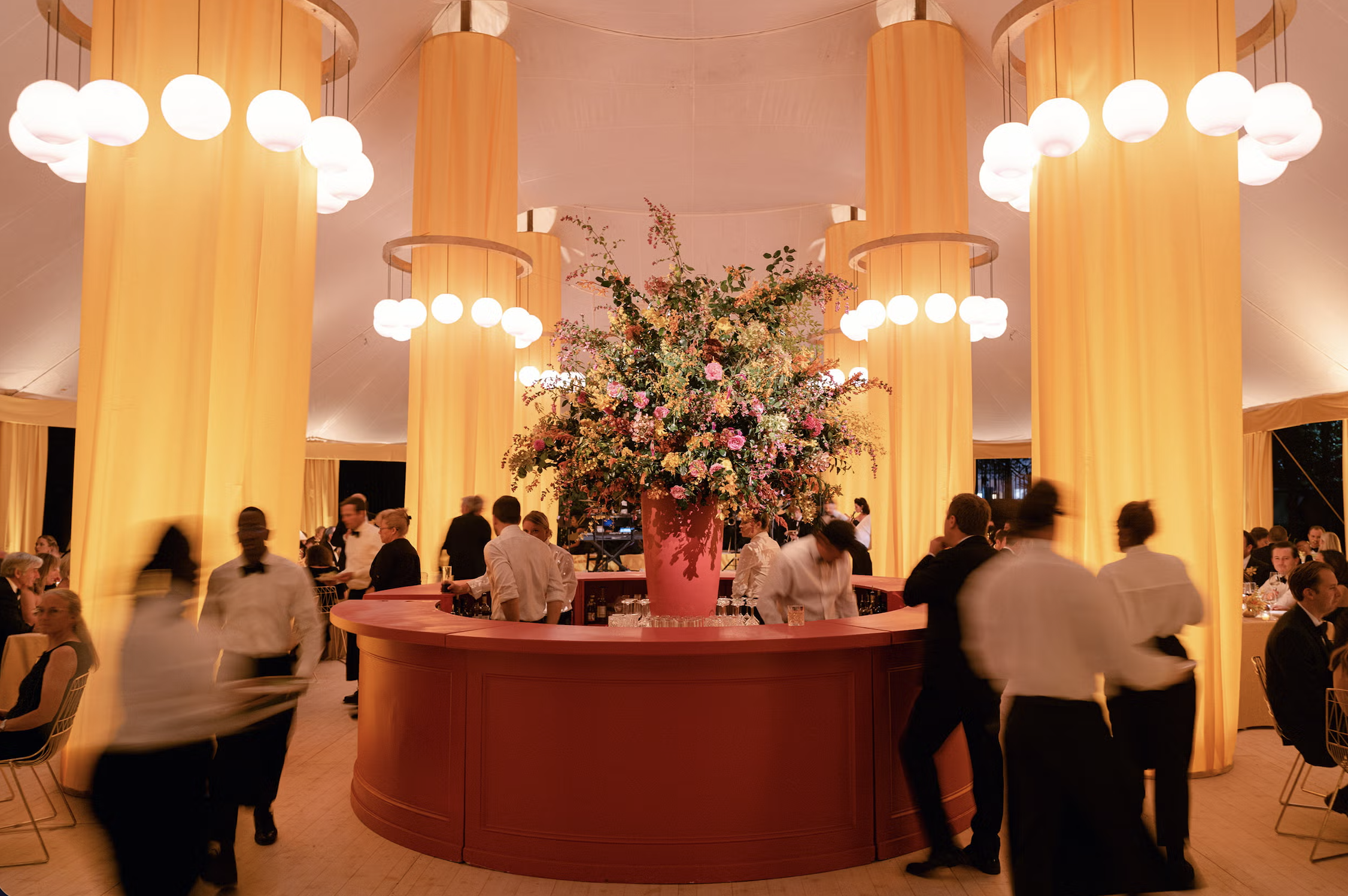 An indoor event with a large floral arrangement on a curved bar in the center, surrounded by people in formal attire, and warm yellow lighting with circular hanging lamps.