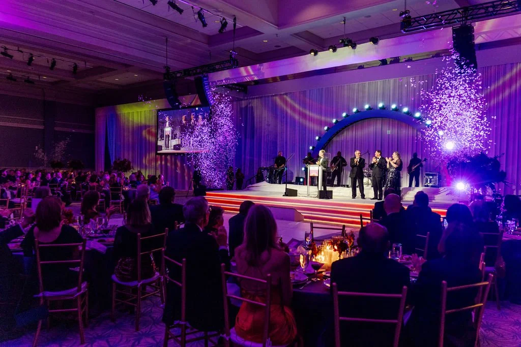 A formal event with a stage performance, decorated with purple and pink lighting, featuring a band singing with their instruments, and an audience sitting at round tables, some with drinks and decorations.