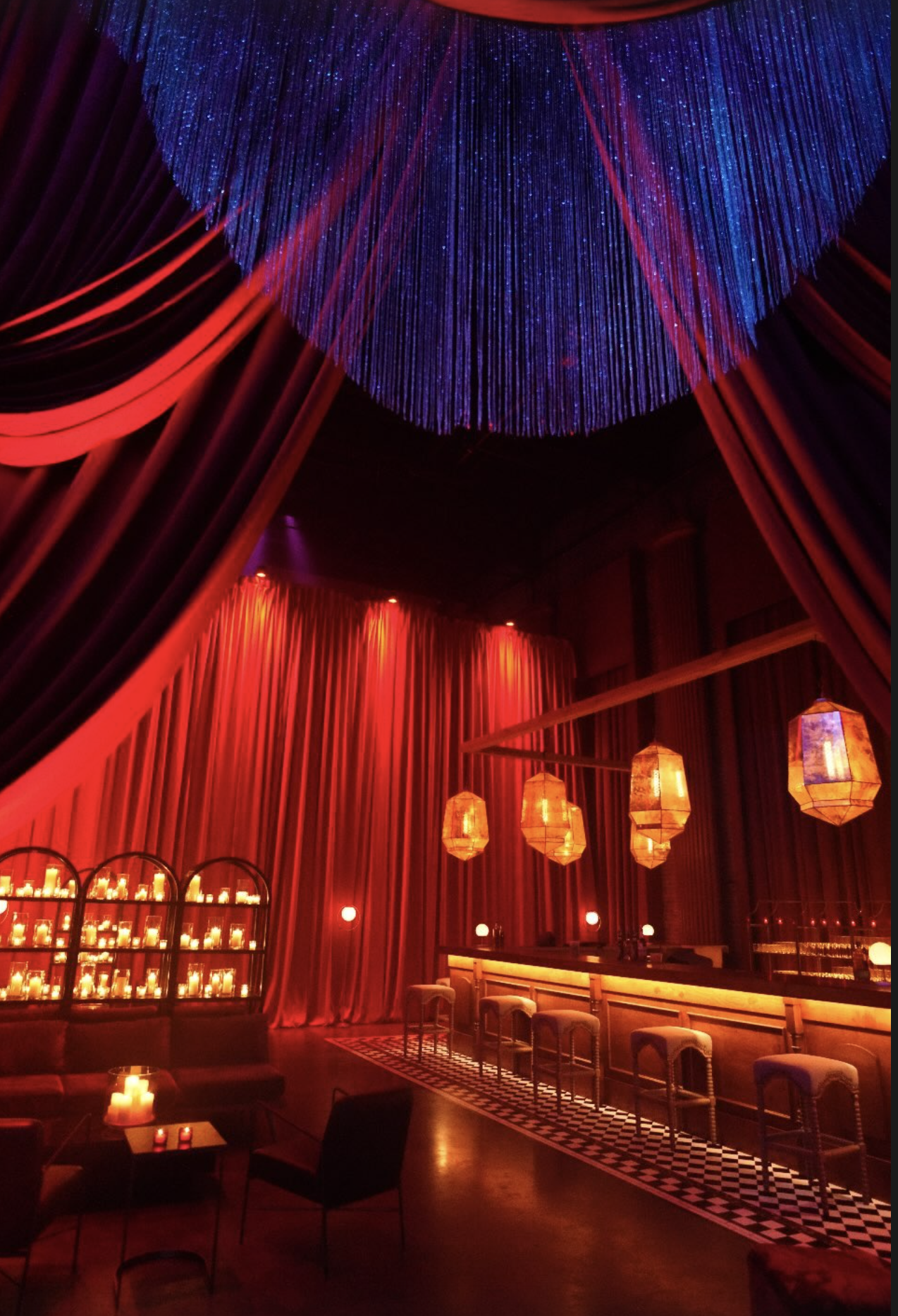 Dimly lit bar or lounge with red curtains, warm lighting, hanging pendant lamps, and a colorful ceiling installation with blue and purple lights.