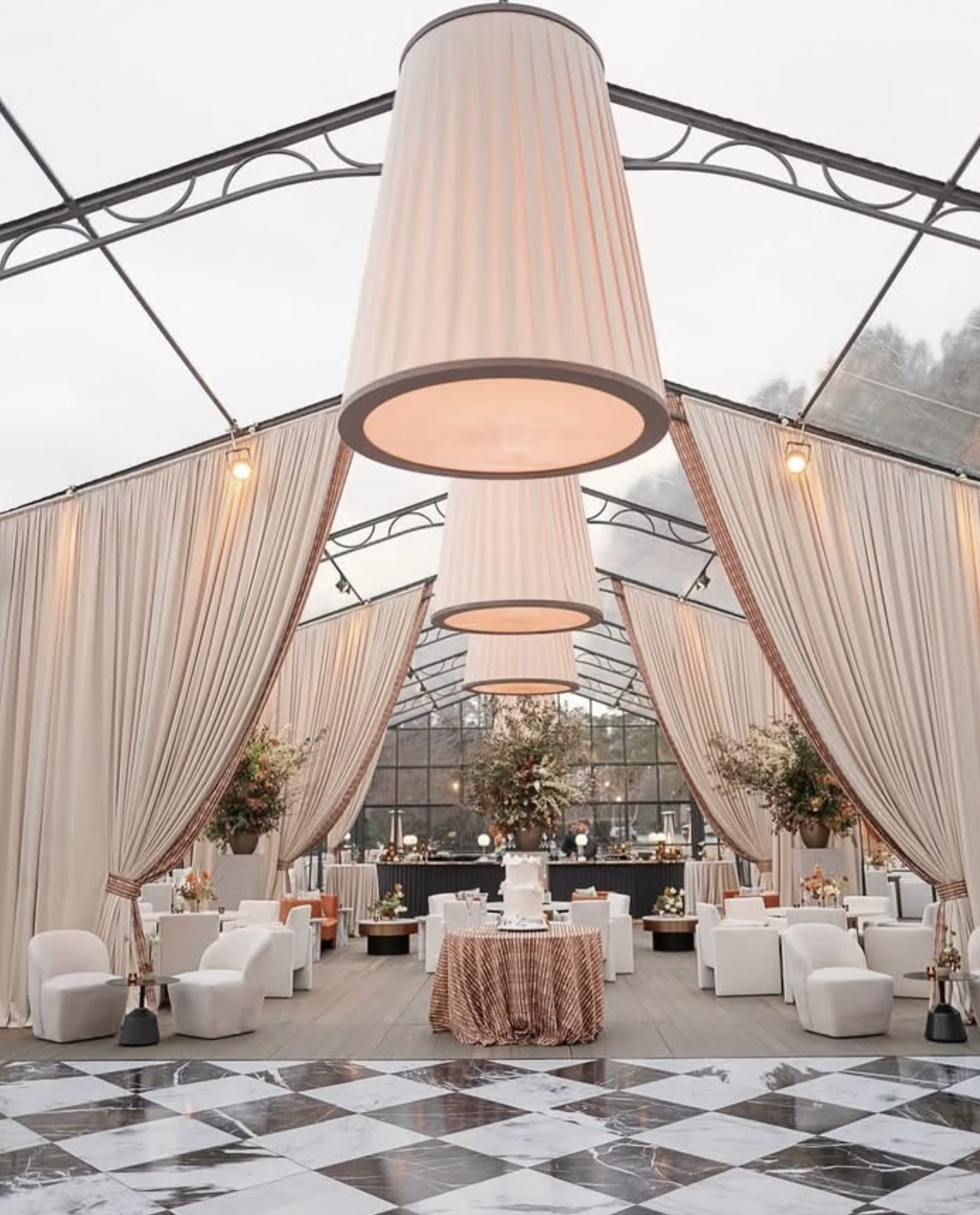 Elegant event space with white chairs, round tables with orange and white tablecloths, large pink and white striped fabric drapes, and a dramatic ceiling with large cylindrical pink and white striped fabric light fixtures hanging from a geometric metal framework. There are floral arrangements and a checkered black and white marble floor.