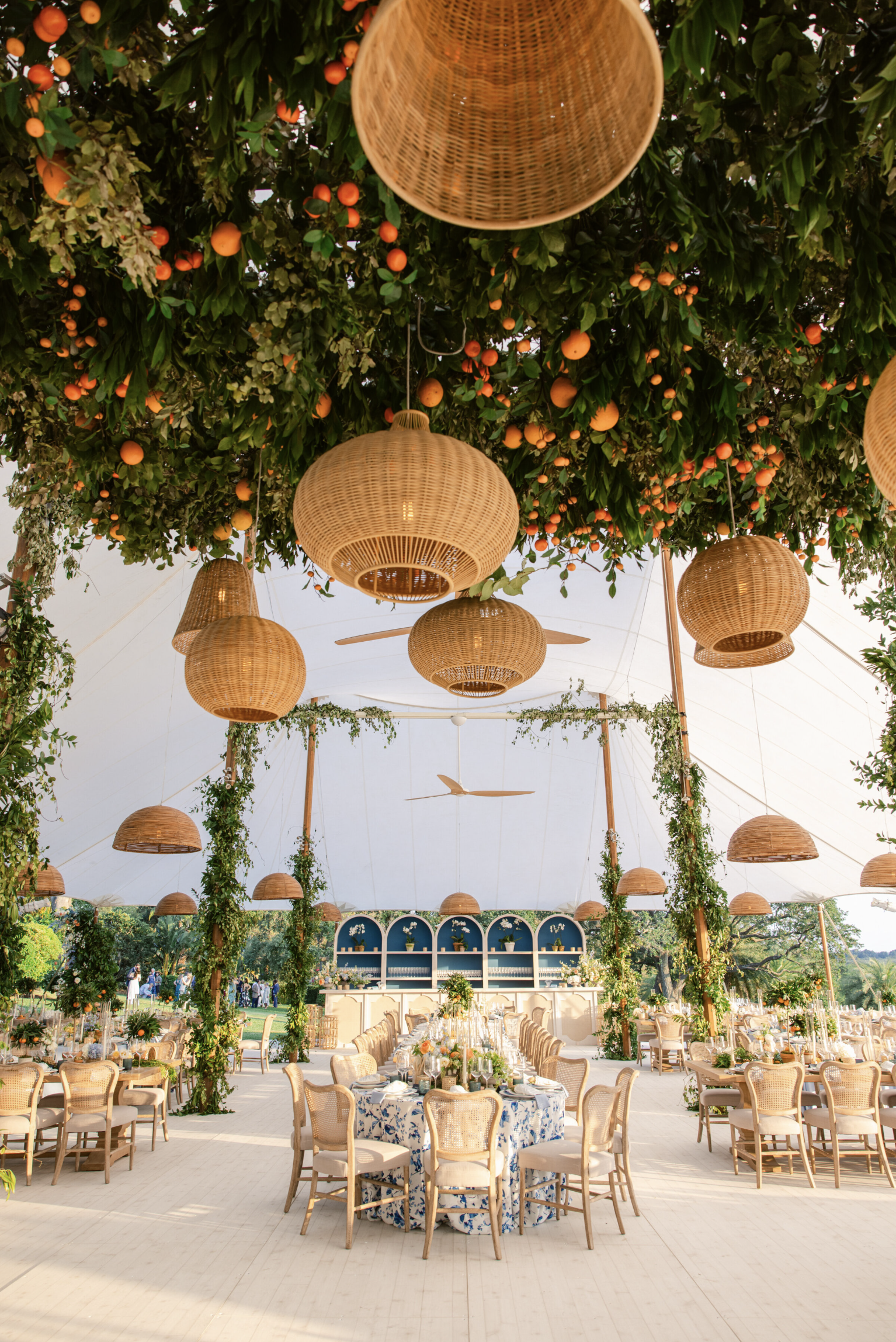 A decorated outdoor event space under a white tent with hanging woven baskets and greenery, set with tables and chairs for a celebration.