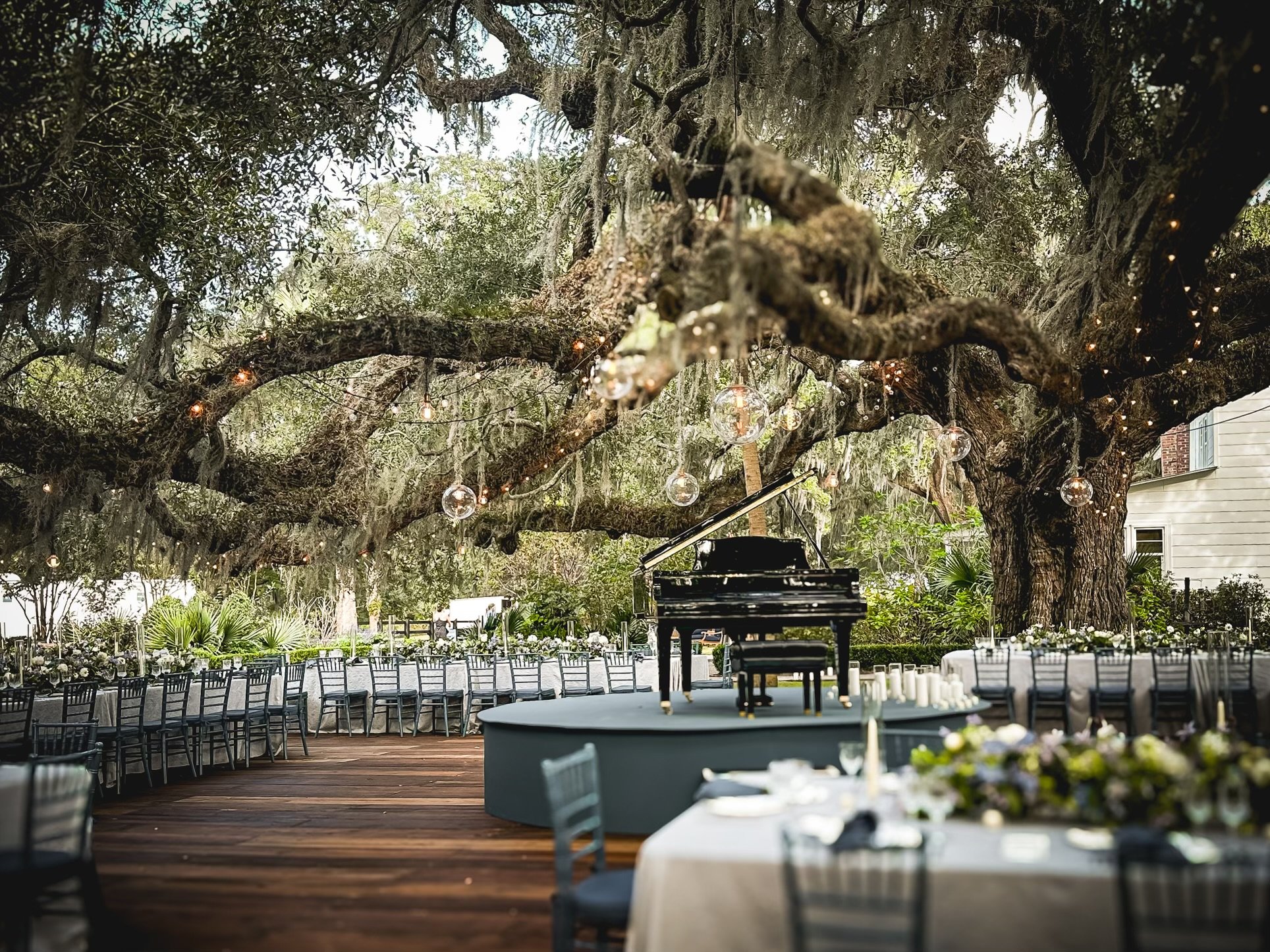 Outdoor wedding setup under large, moss-draped trees with string lights, chairs arranged facing a round stage, and a grand piano in the center.