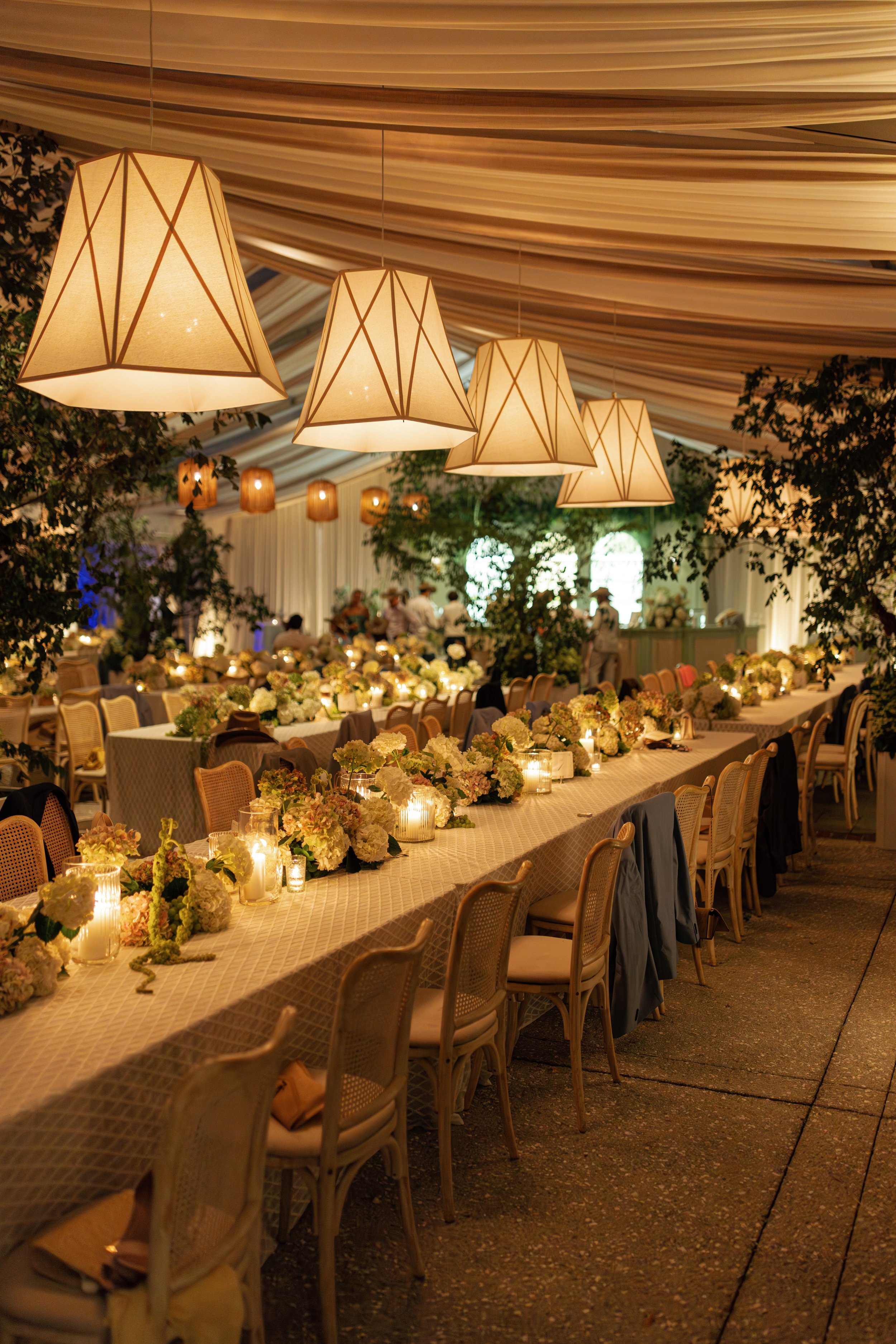 Elegant event setup with long tables decorated with white and pastel flowers, candles, and tea light holders. Large beige pendant lights with geometric patterns hang from a ceiling draped with white fabric. In the background, there are guests, greenery, and additional lighting creating a warm ambiance.