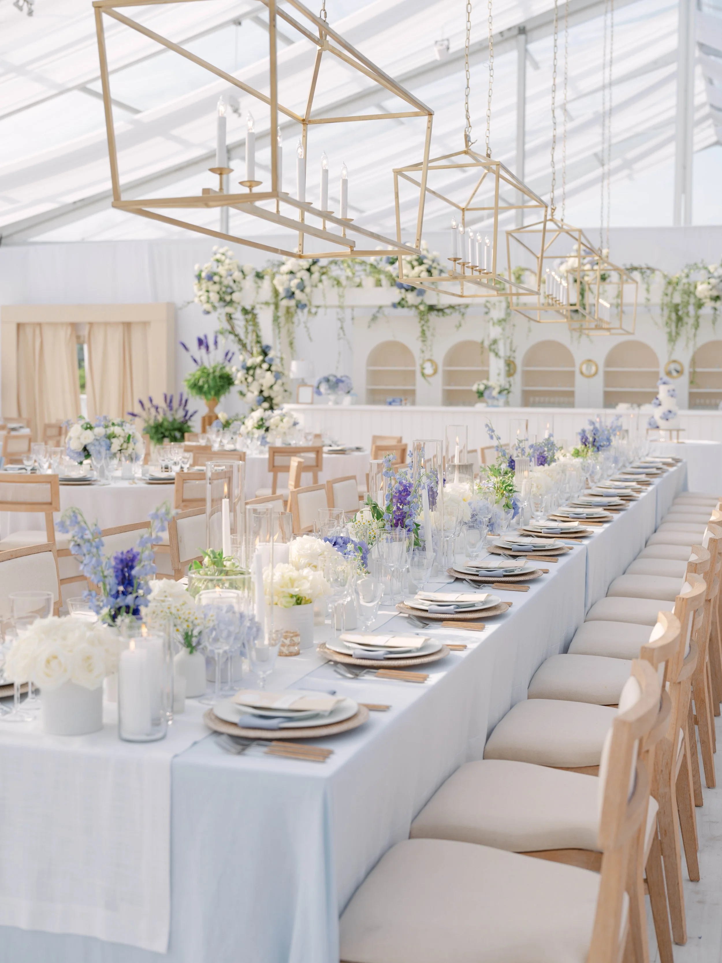 Elegant wedding reception setup featuring long white tables with floral centerpieces, gold chandeliers hanging from the ceiling, and cream-colored chairs.