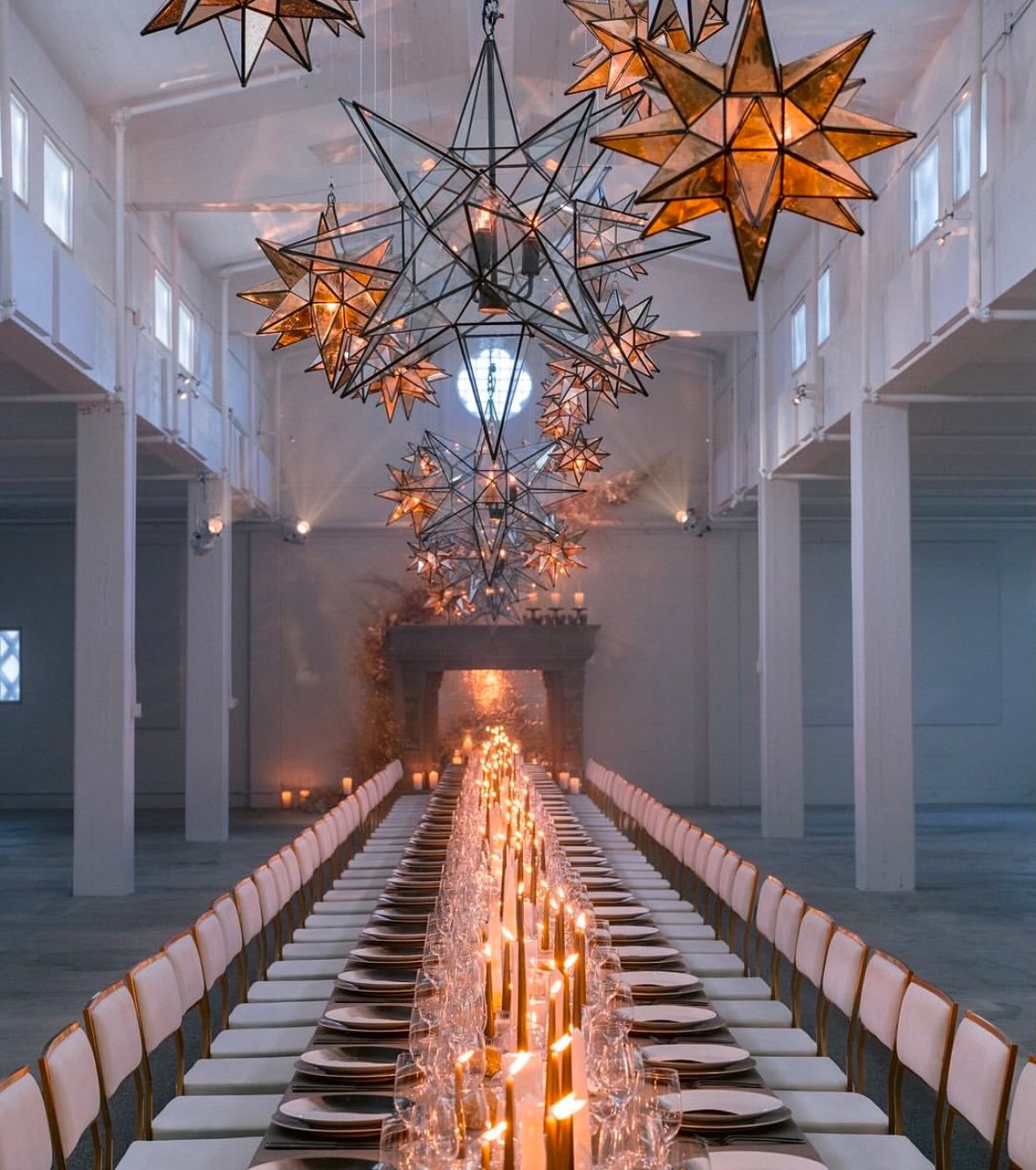 Long dining table set with plates, glasses, napkins, and lit candles, leading to a fireplace with candles and a floral arrangement, under large star-shaped pendant lights hanging from the ceiling of a spacious, white-walled hall.