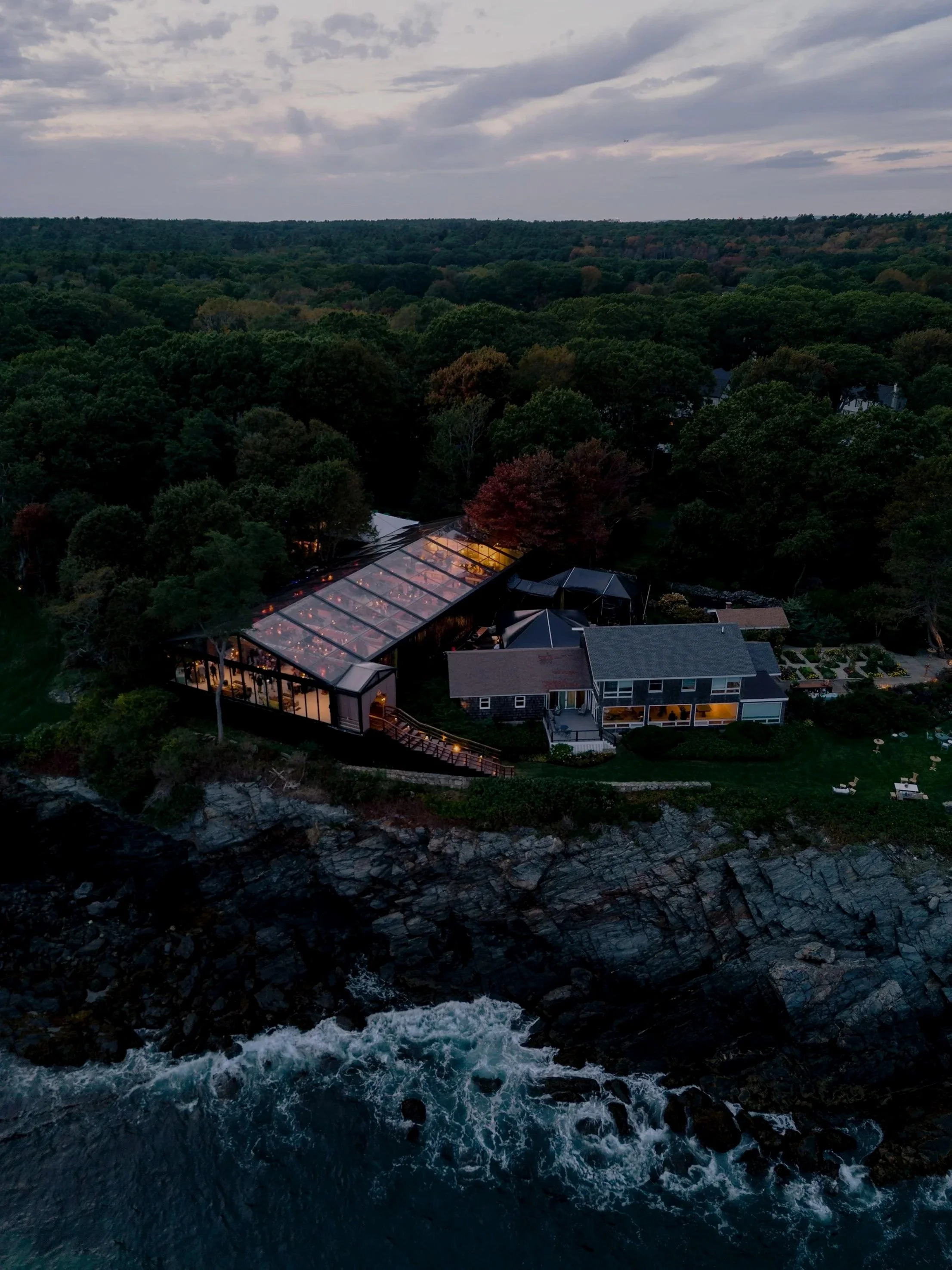 An aerial night view of a house with a glass roof in a wooded area near the shoreline, with water and rocks in the foreground and cloudy sky above.
