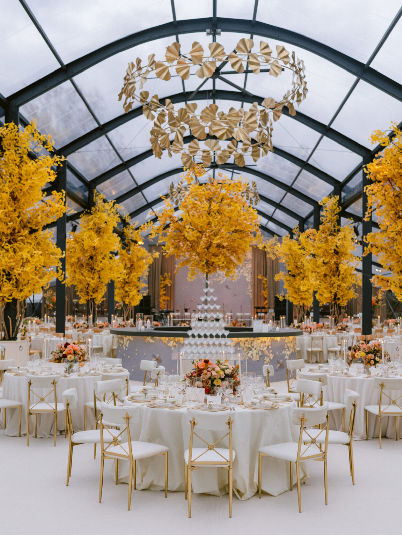 Elegant event hall decorated with large yellow floral arrangements, round tables with white tablecloths and gold accents, and a modern chandelier hanging from a glass arched ceiling.