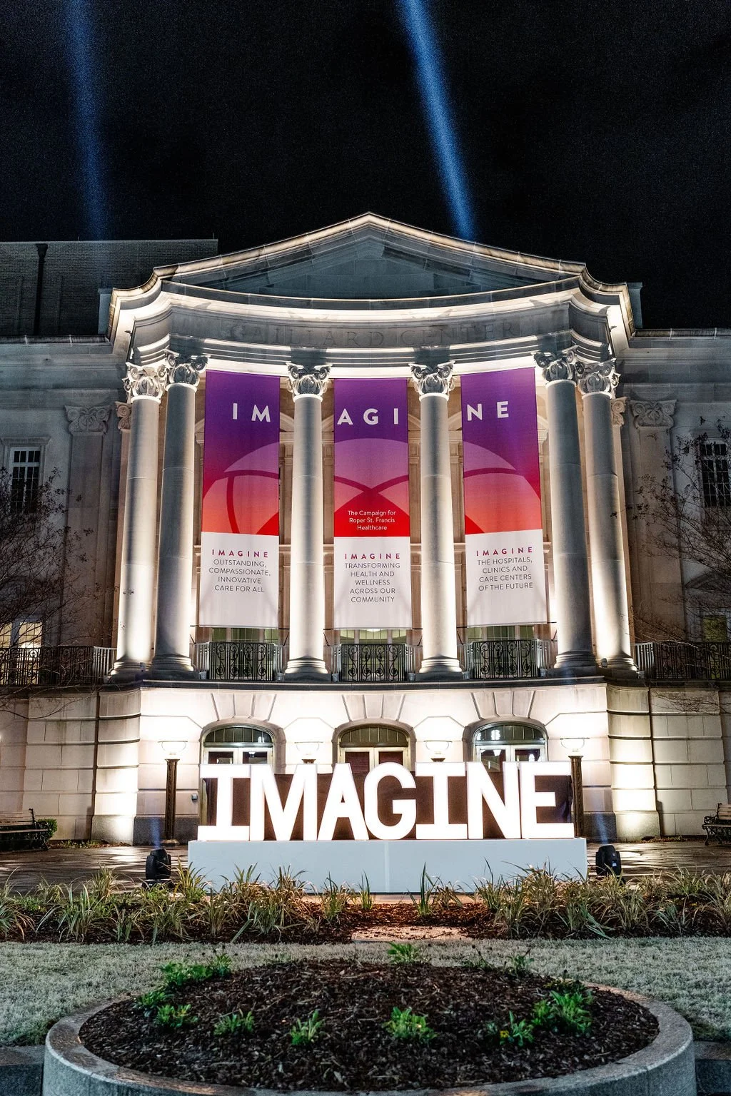 Night view of a classical building with large illuminated white sign that spells 'IMAGINE' in front, and three banners reading 'IMAGINE' hanging from columns, promoting healthcare innovation and community wellness.