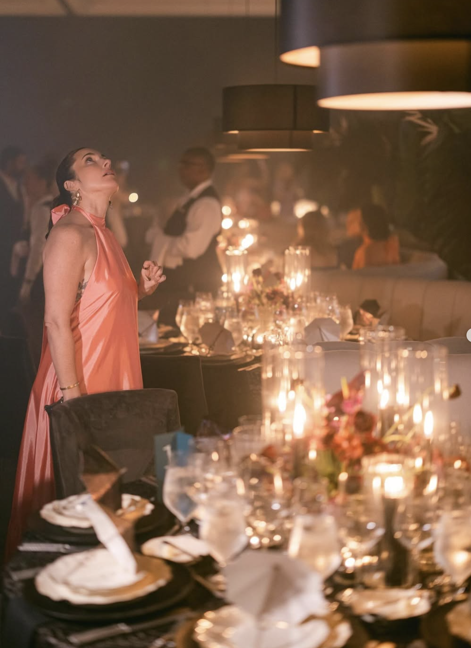 A woman wearing a pink satin dress looking up in a dimly lit restaurant or lounge with hanging pendant lights and tables decorated with floral centerpieces and candles.