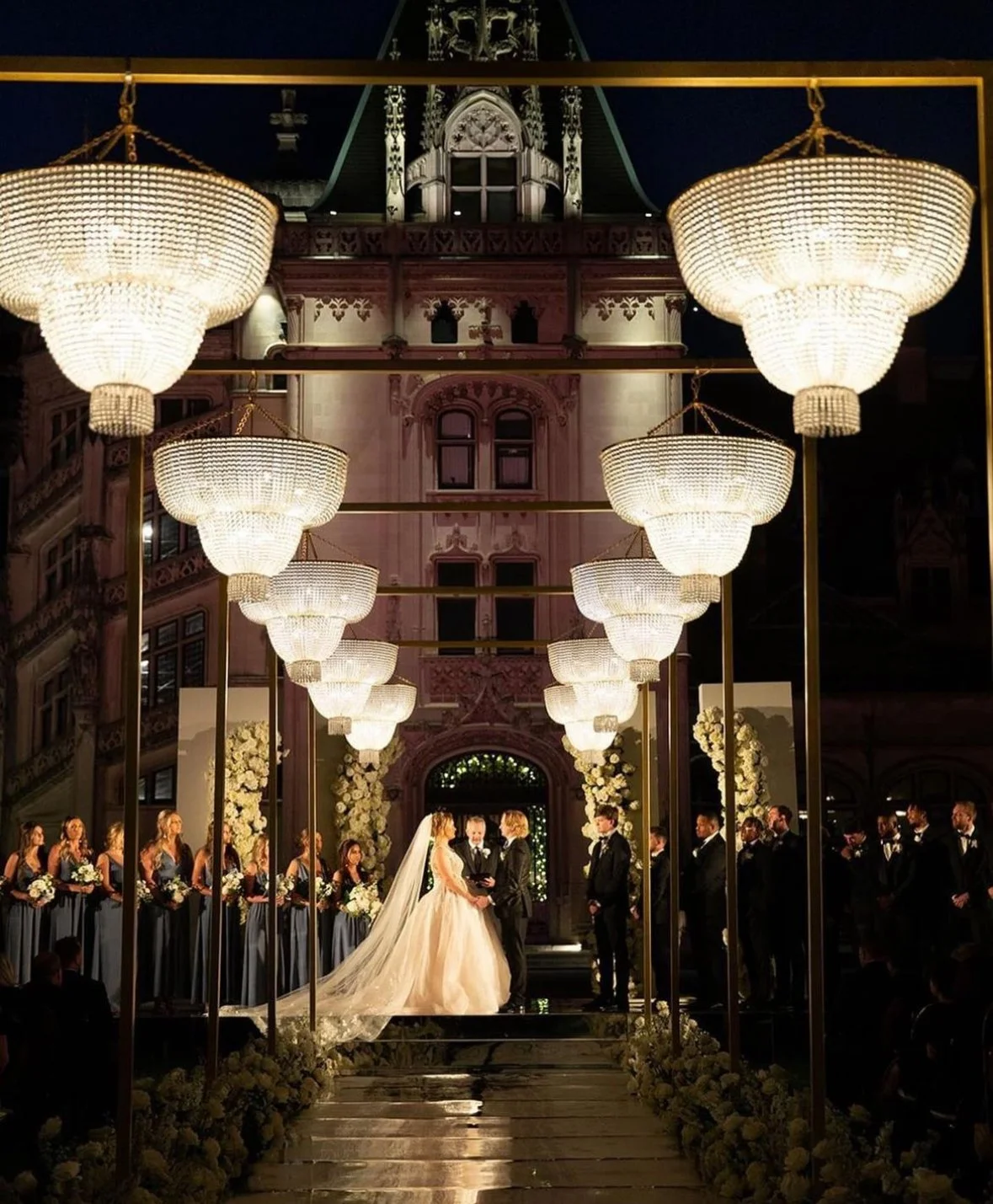 A wedding ceremony taking place outdoors at night under large hanging chandeliers, with a bride and groom exchanging vows surrounded by bridesmaids and groomsmen, in front of an ornate historic building.