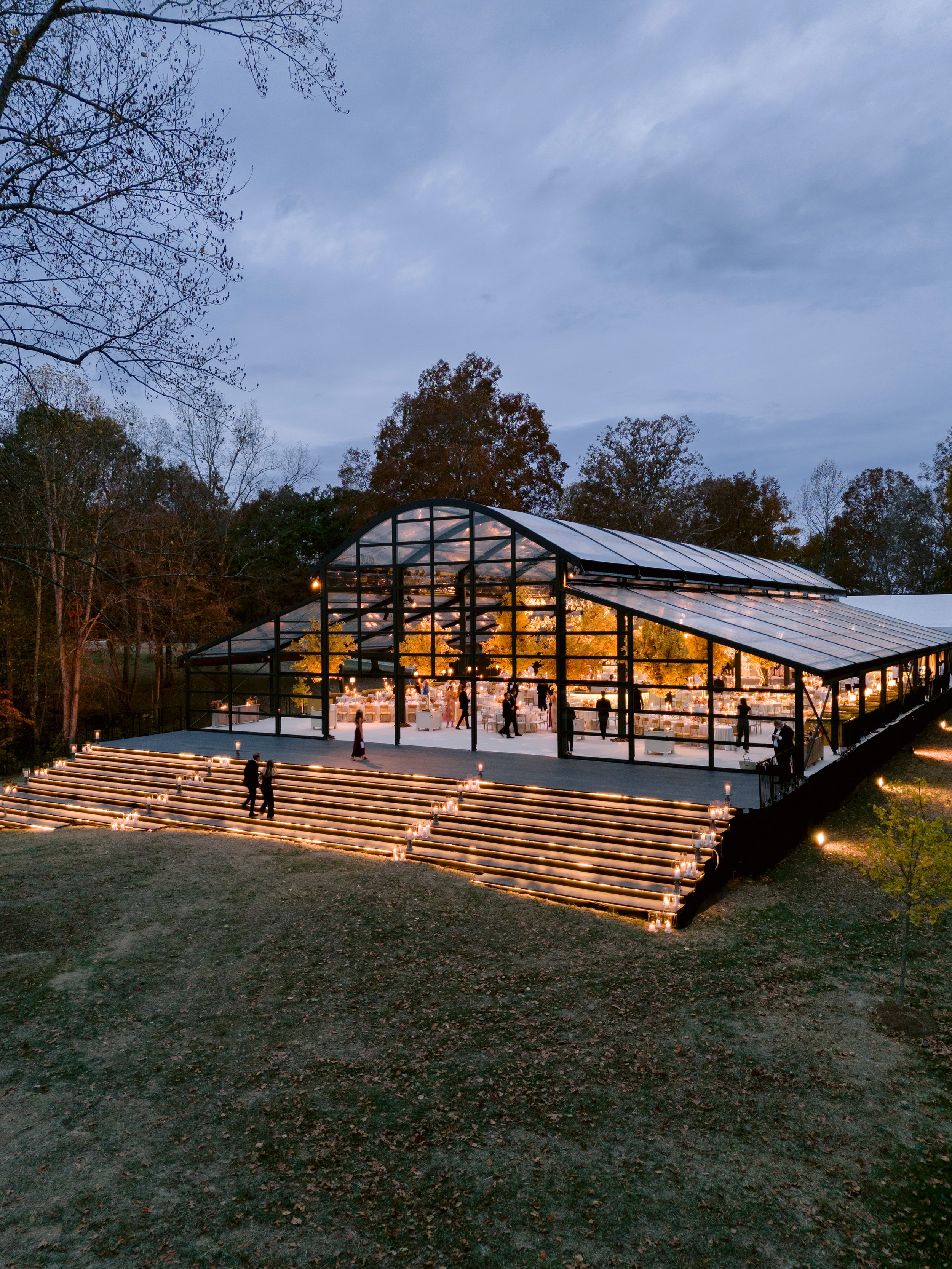 A glass greenhouse illuminated from within, set outdoors at dusk, with trees and a cloudy sky in the background. Guests are mingling inside and on the outdoor steps lined with candles.