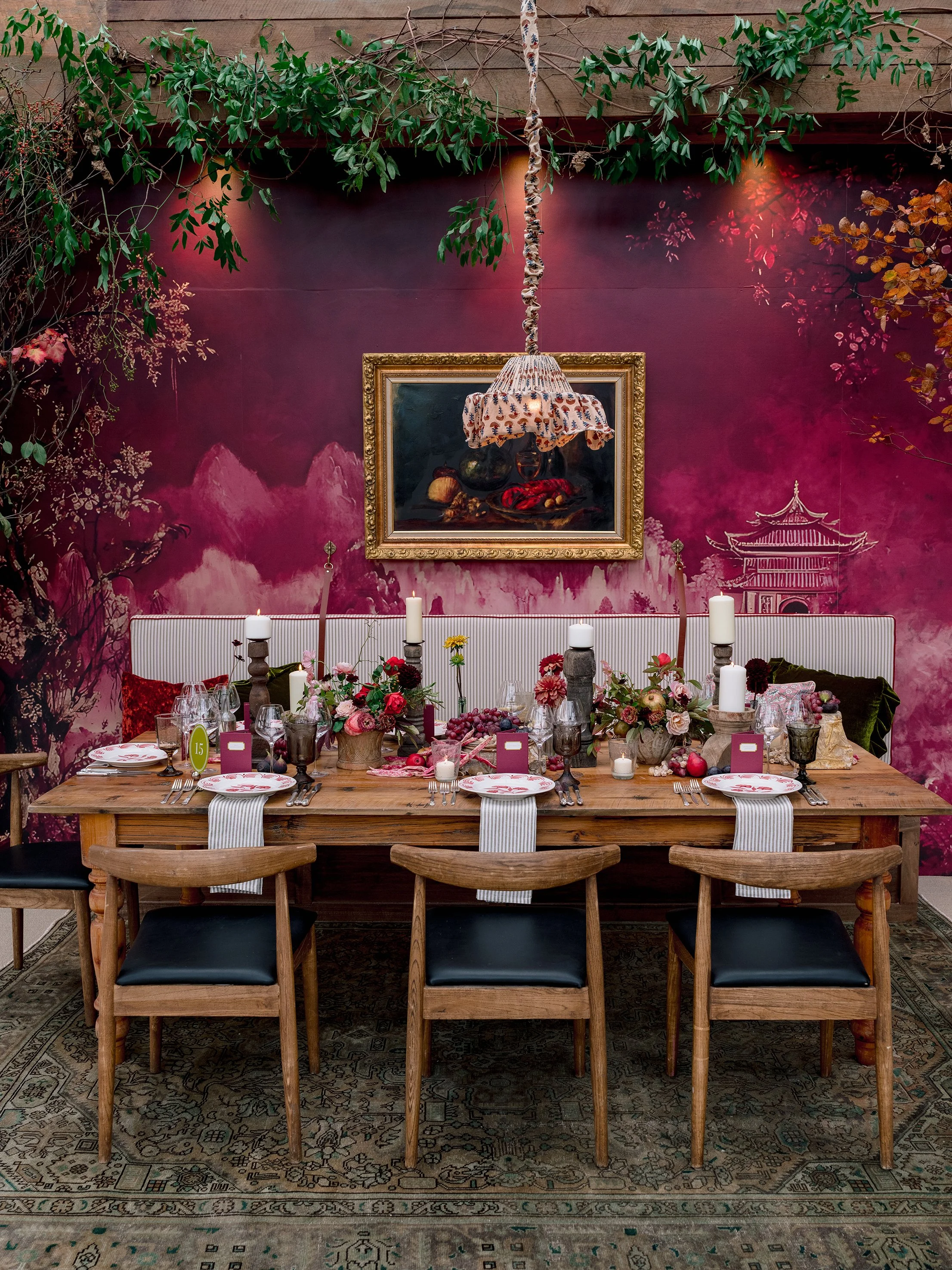 A dining table set for a meal with floral arrangements, lit candles, and place settings. The room has a pink mural with mountain and pagoda artwork, greenery, and a painting of a still life with fruit and a cloth lamp shade hanging above.