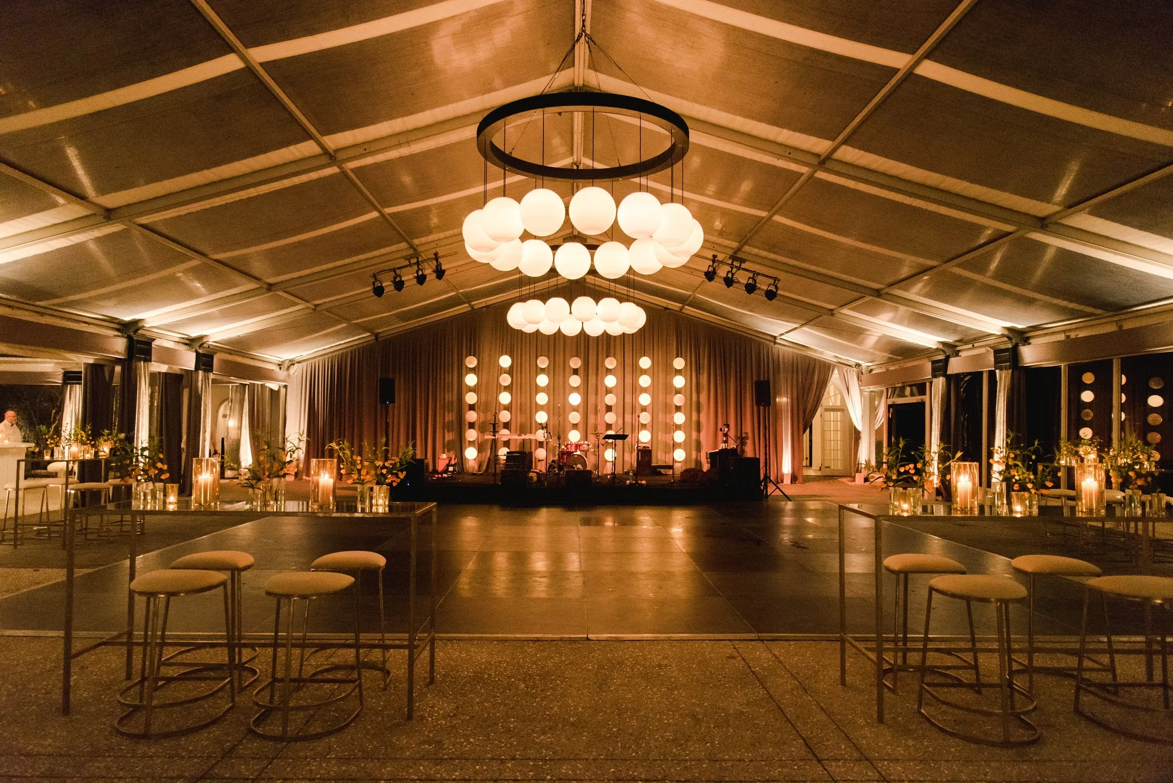 Elegant indoor event venue with a stage set up for a performance, large circular chandelier with multiple round lights hanging from the ceiling, surrounded by tables and chairs with candles and floral arrangements, beige curtains, and multiple string lights on the walls.