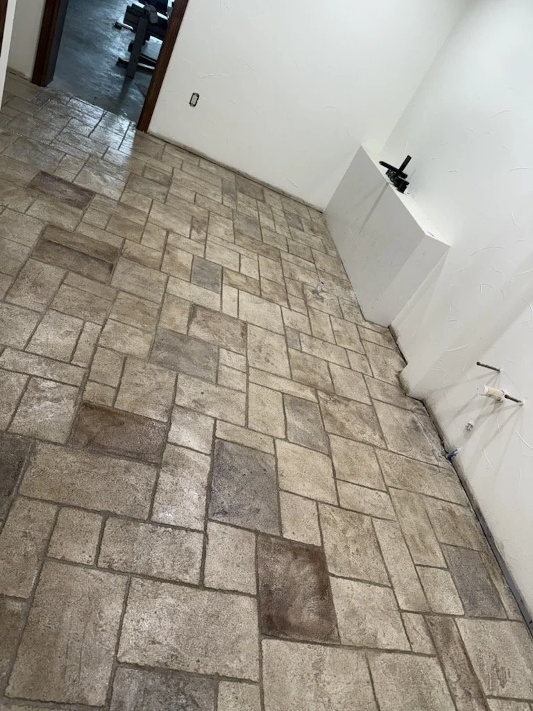 Interior room with tiled stone floor, white walls, and an opening to another room with exercise equipment. There is a white stand or pedestal in the corner with some tools or objects on top, and exposed pipes or wires on the wall.