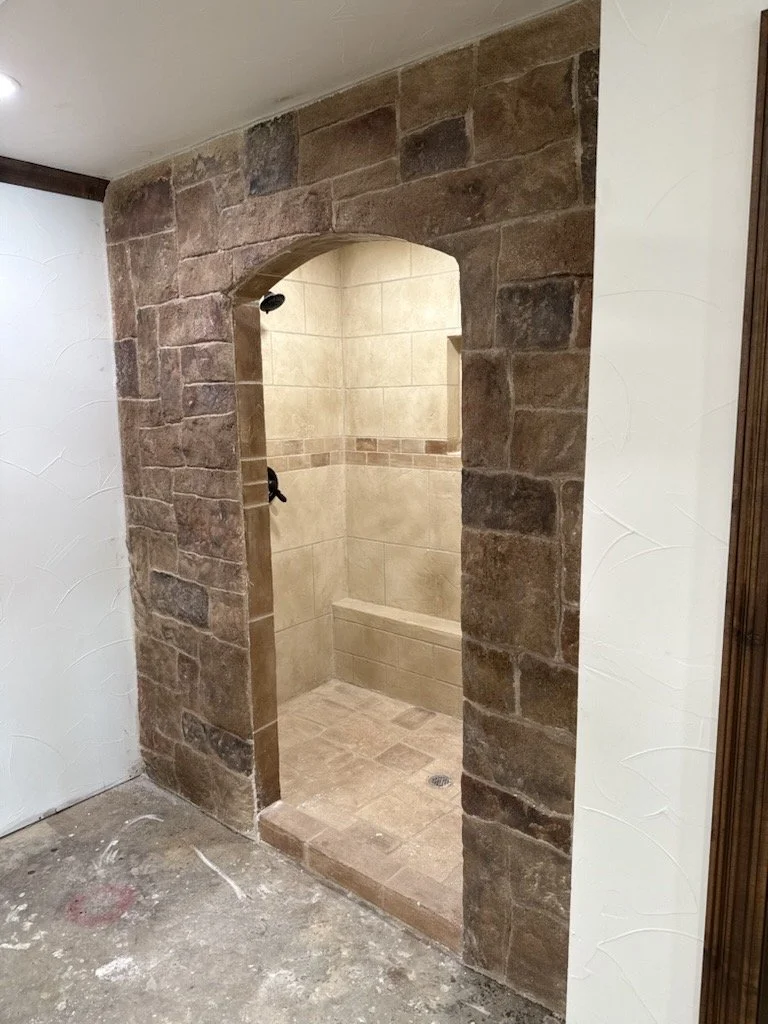 Inside a shower with beige tiles, a built-in bench, and a dark shower head, surrounded by a brick archway and wall.