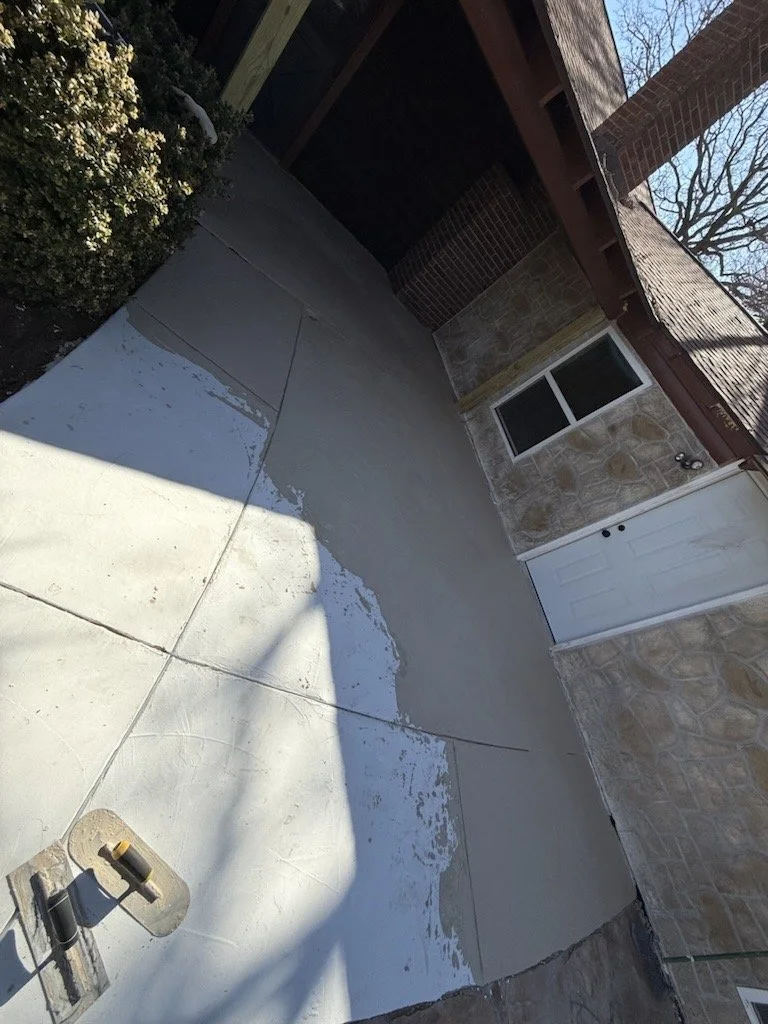 Partially concrete porch with some areas freshly painted or coated, adjacent to stone and brick house exterior, with a window and door, and a skateboard lying on the ground.