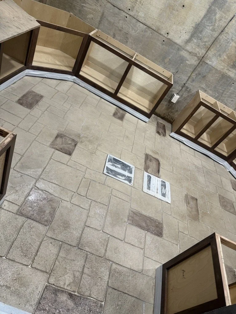Empty wooden display cases arranged in a U-shape on a concrete floor with a concrete wall in the background. Two ventilation covers are visible on the floor.