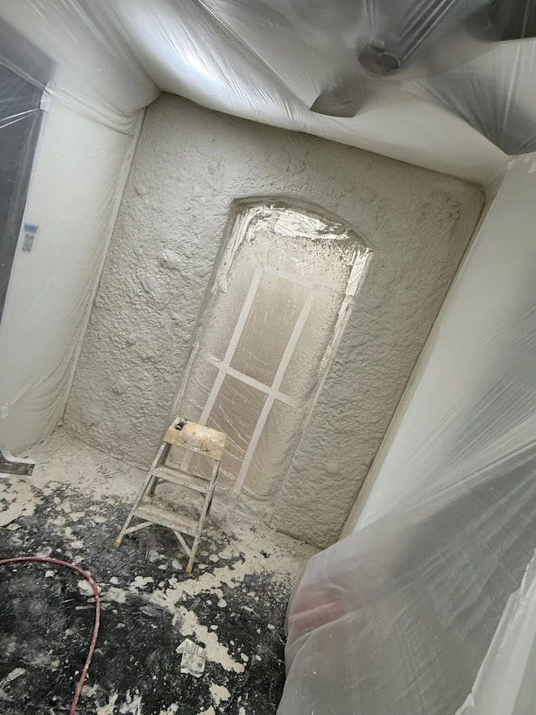 A construction or renovation room with a window being coated in spray texture, plastic sheeting covering the walls and ceiling, a small wooden step ladder in the center, and debris on the dark floor.
