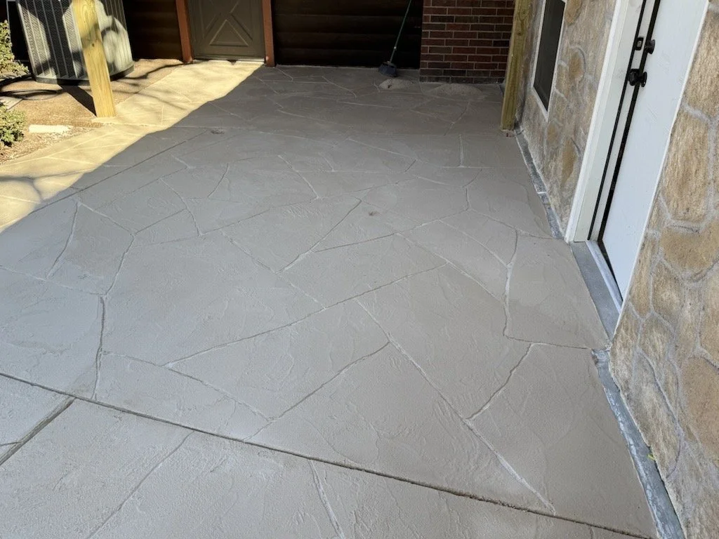 Concrete patio with decorative stone pattern adjacent to house with brick and stone exterior, door, and window.