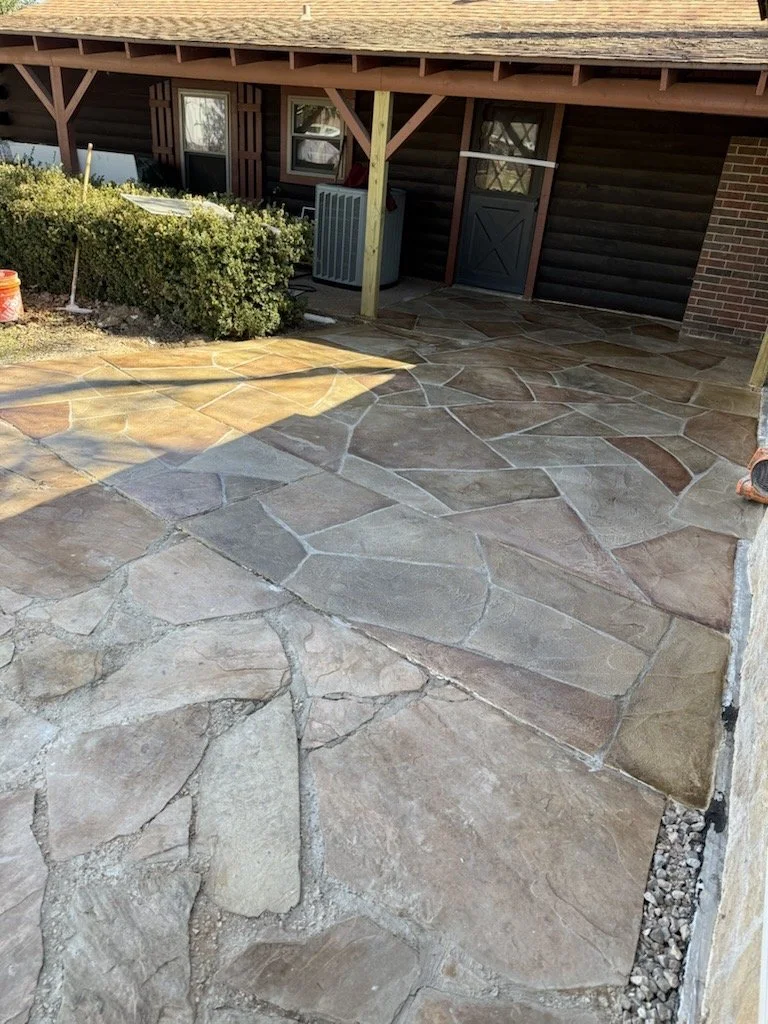 A stone patio with irregularly shaped, multicolored stones leading up to a house with a covered porch, bushes, and an air conditioning unit.