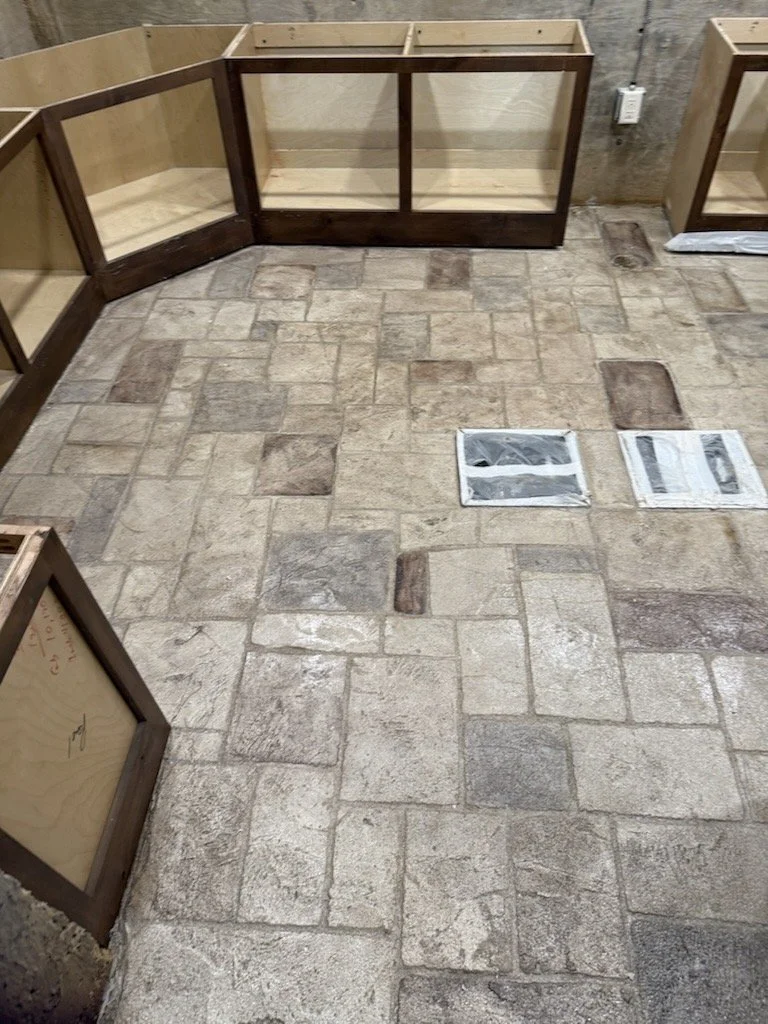 Room with partially assembled wooden cabinets and a tiled floor with two floor vents.