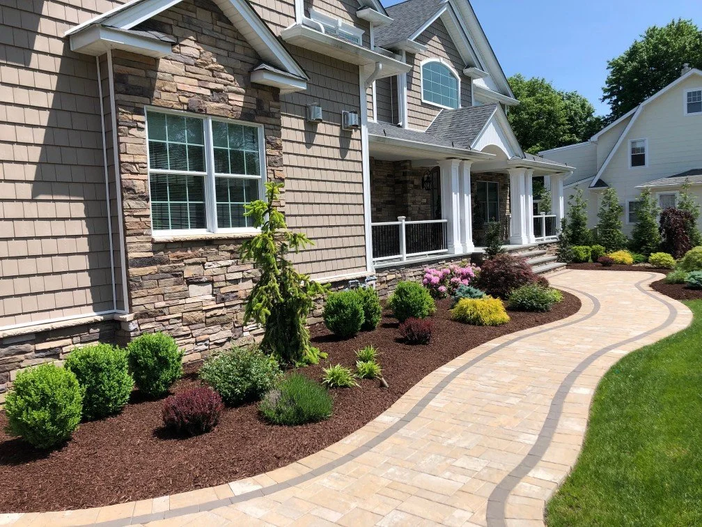 A house with a front yard featuring a curved brick walkway, lush green grass, and well-maintained landscaping with various bushes and small trees.