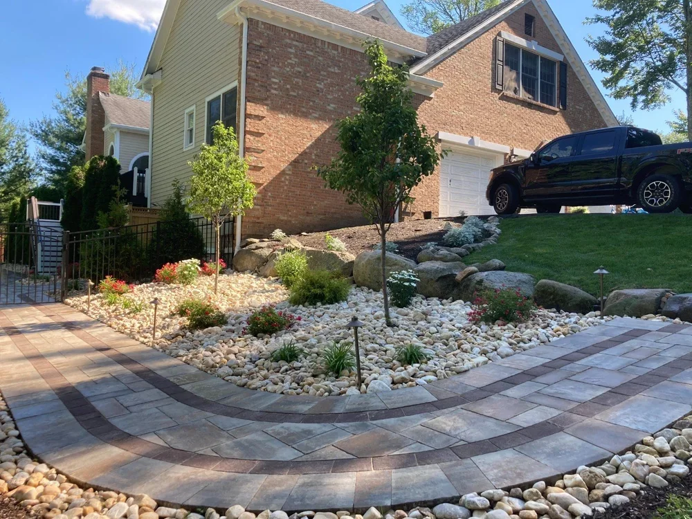 Front yard with landscaped garden featuring rocks, small bushes, and trees, alongside a paved walkway leading to the house. The house has a brick and siding exterior with a garage and a black vehicle parked outside.