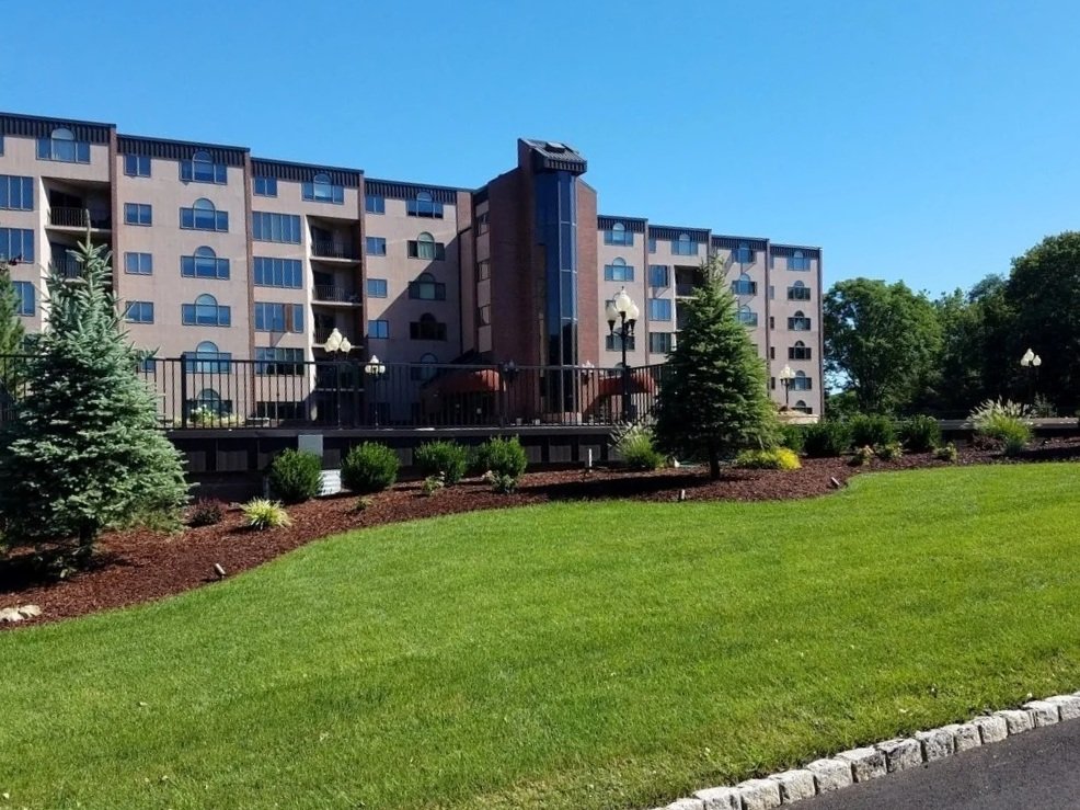 Multi-story residential building with brick facade, manicured green lawn, decorative landscaping beds with mulch, and evergreen trees along the entrance - GreenNature Landscape Design